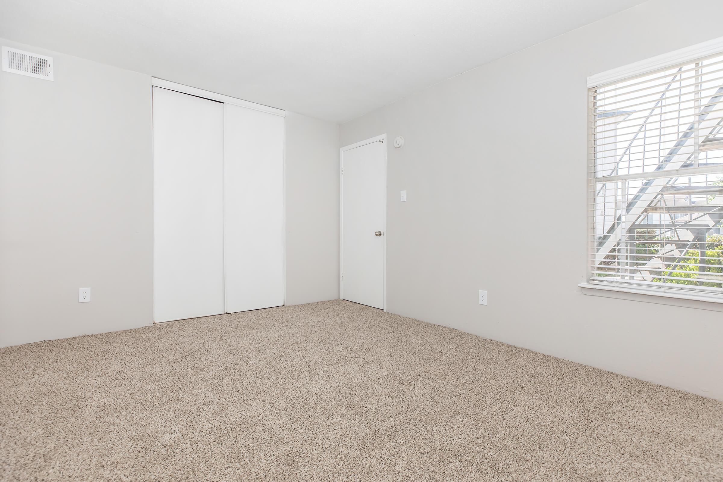 Empty room with light grey walls and beige carpet. There is a closet with sliding doors on the left, a white door on the right, and a window with blinds allowing natural light to enter. The overall atmosphere is bright and minimalistic, suitable for various uses.