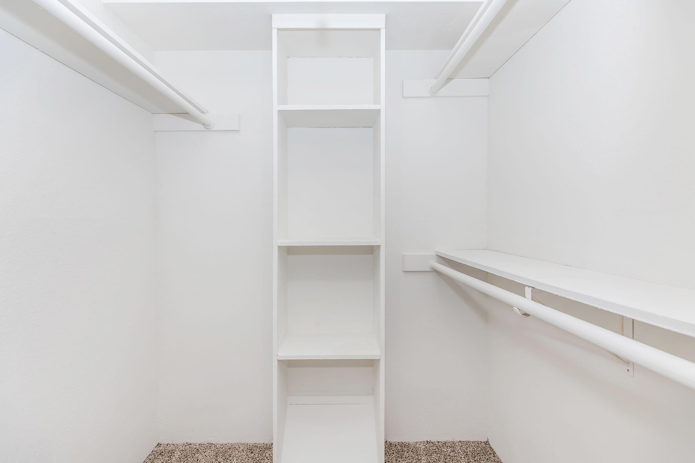 A clean, empty closet with white walls and shelves. There are vertical shelves in the center and two horizontal rods for hanging clothes on either side. The floor is carpeted with a light texture, creating a minimalist and organized storage space.