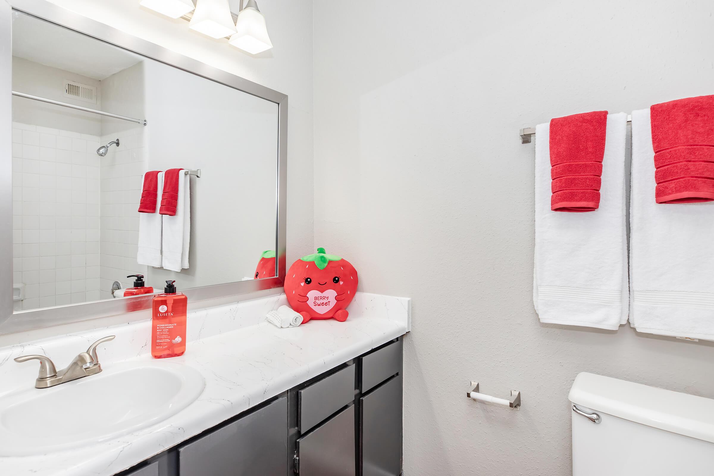 A clean bathroom featuring a white sink with a silver faucet, a large mirror above it, and neatly arranged red towels hanging on a rack. A decorative strawberry-shaped plush toy sits beside the sink, adding a playful touch. The walls are light-colored, and the overall decor is bright and inviting.