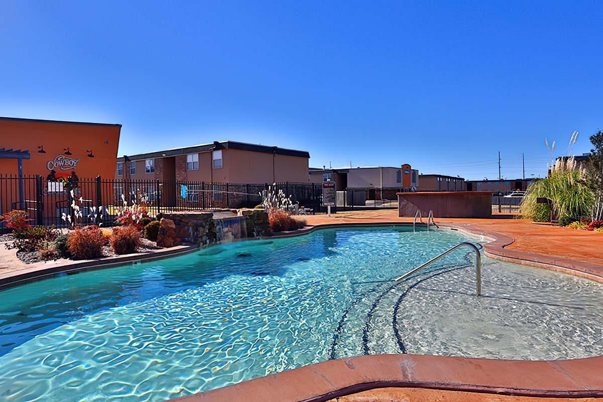 A clear swimming pool with a gentle waterfall, surrounded by lush greenery and colorful flowers. In the background, there are several buildings against a bright blue sky. The scene is inviting and perfect for relaxation.