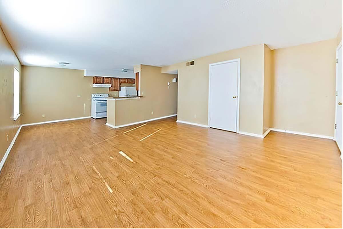 A spacious, empty living area with light brown walls and hardwood flooring. The kitchen is partially visible in the background, featuring a stove and cabinetry. Natural light enters through the windows, highlighting the open layout and clean, uncluttered space.