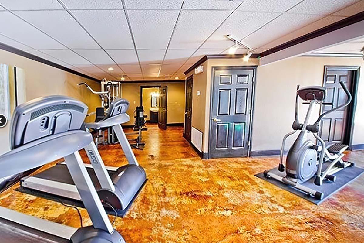 A view of an indoor gym featuring two treadmills and an elliptical machine. The room has a modern design with a polished floor and well-lit areas. There are doors leading to other spaces in the background, creating an inviting workout environment.