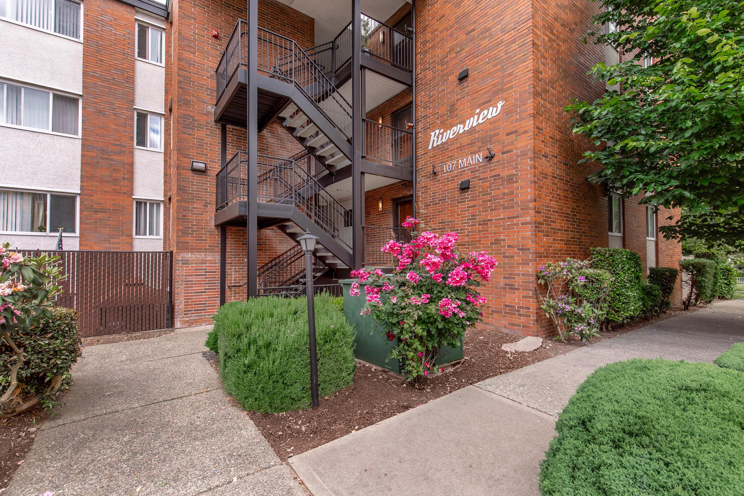 A brick apartment building with a prominent staircase leading to an upper entrance. Green shrubs and colorful pink flowers are planted alongside the walkway, which is paved and leads from the street to the entrance. The building features a modern design with multiple windows.
