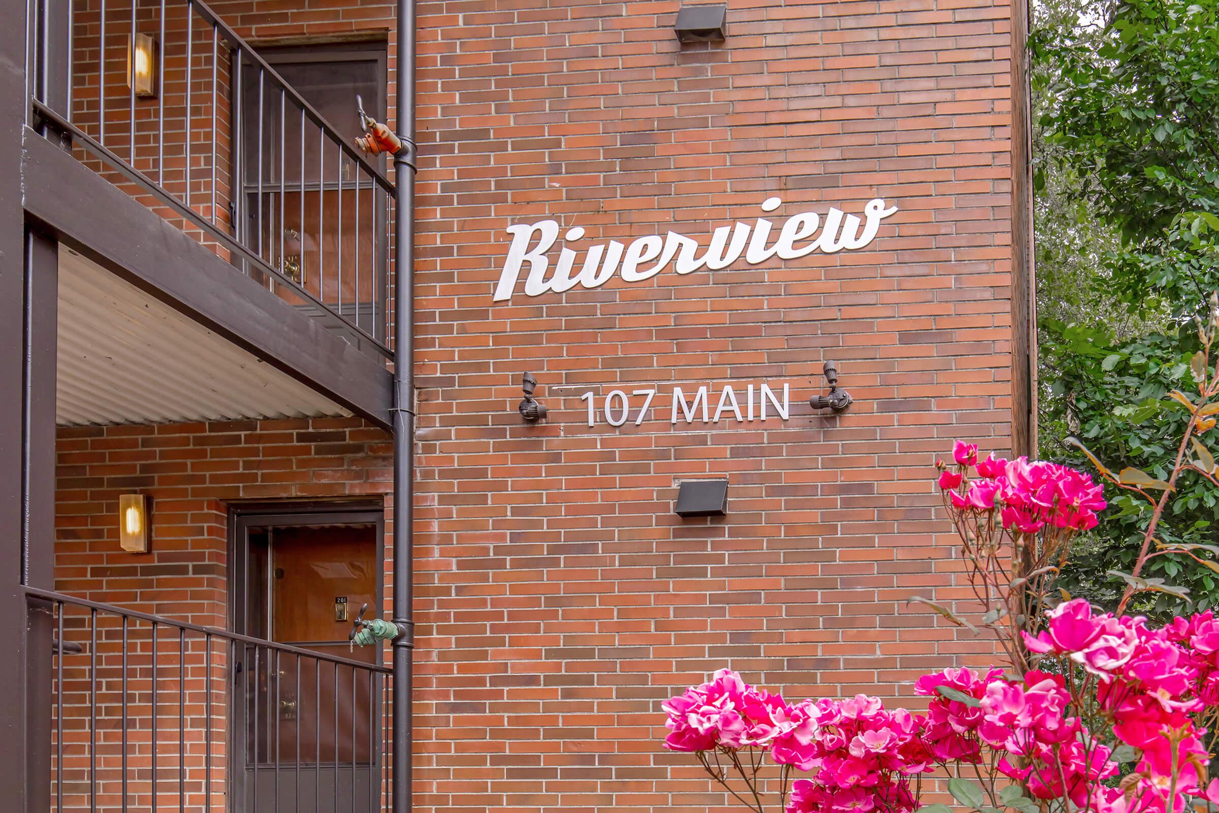 Sign on a brick building reading "Riverview 107 MAIN," accompanied by colorful pink flowers in the foreground. The building features a balcony with black railings and a warm atmosphere.