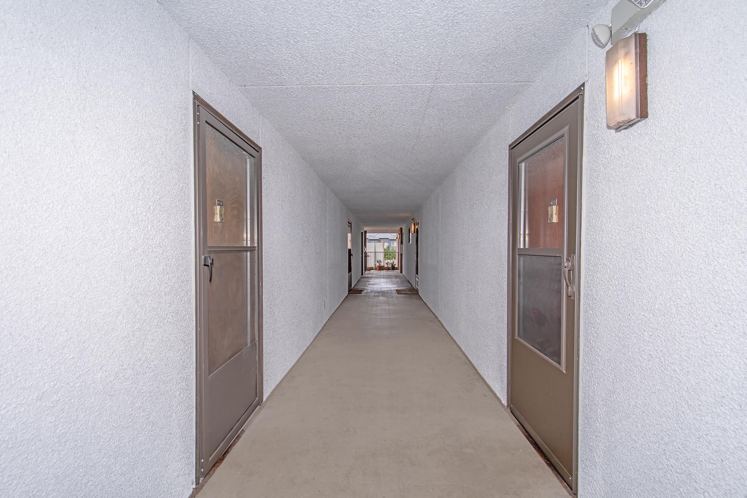A well-lit corridor featuring two sets of doors on either side, with a smooth concrete floor and textured white walls. At the far end, light floods in from an open space, creating a sense of depth and inviting atmosphere. Wall-mounted lights provide additional illumination along the hallway.