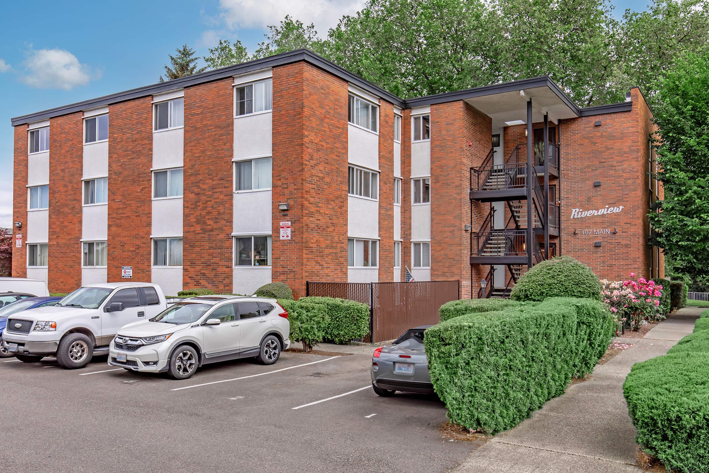 A three-story brick apartment building named "Riverview" surrounded by greenery. The structure features multiple windows, a staircase at the side, and is accompanied by a parking lot with several cars. The scene is set against a backdrop of trees and a cloudy sky.