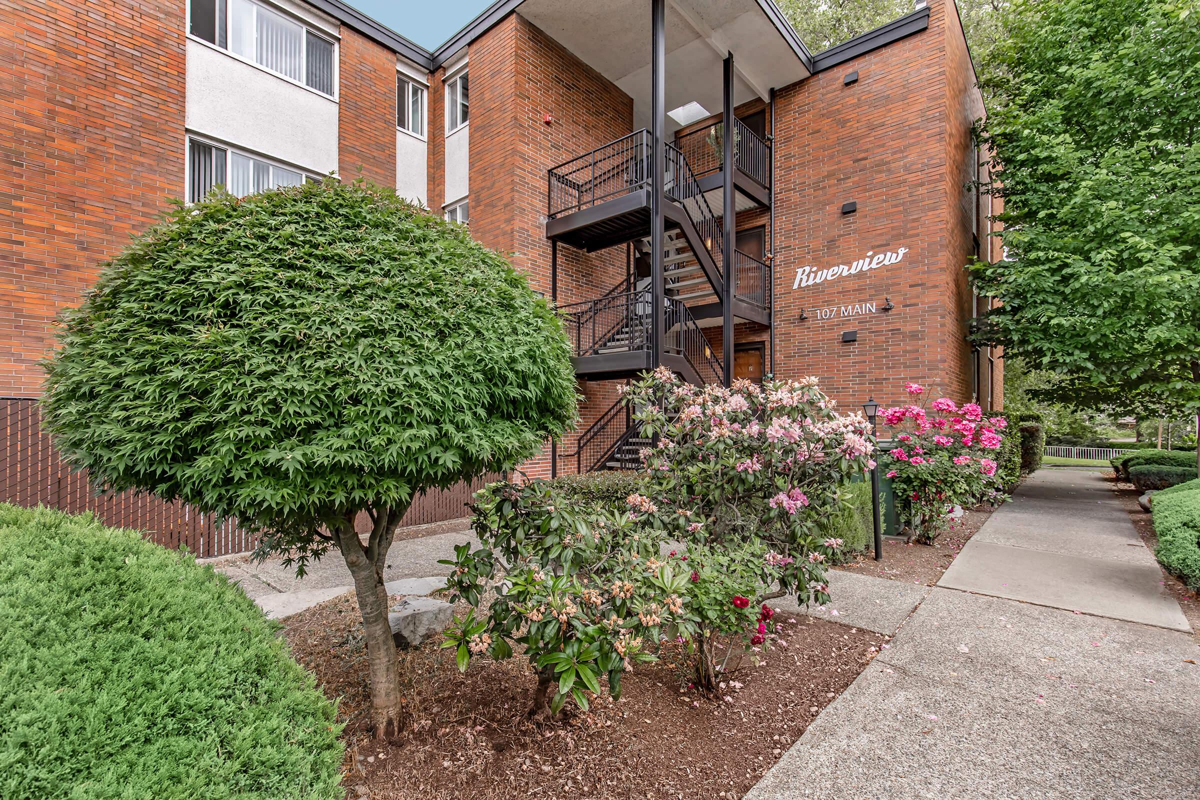 Exterior view of a three-story brick apartment building named "Riverview." The image features neatly trimmed shrubs and vibrant flower beds with pink blooms in the foreground. A staircase leads to the upper floors, and trees line the walkway, creating a pleasant and inviting atmosphere.
