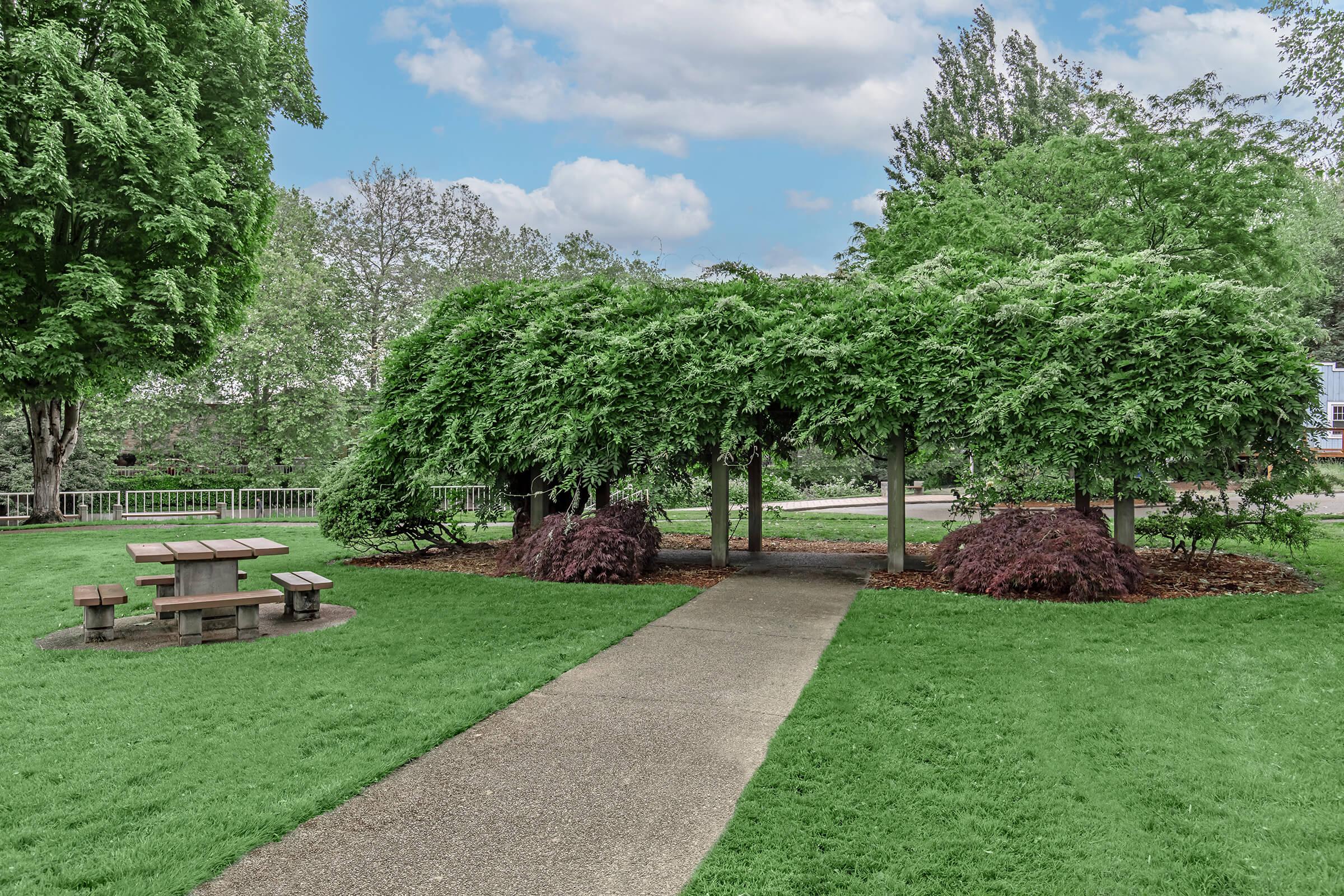 A landscaped park featuring a path leading to a canopy of lush green foliage. To the left, there is a wooden picnic table under the trees, and surrounding the area are various shrubs and vibrant greenery, creating a serene outdoor space ideal for relaxation. The sky is partly cloudy, adding to the tranquil atmosphere.