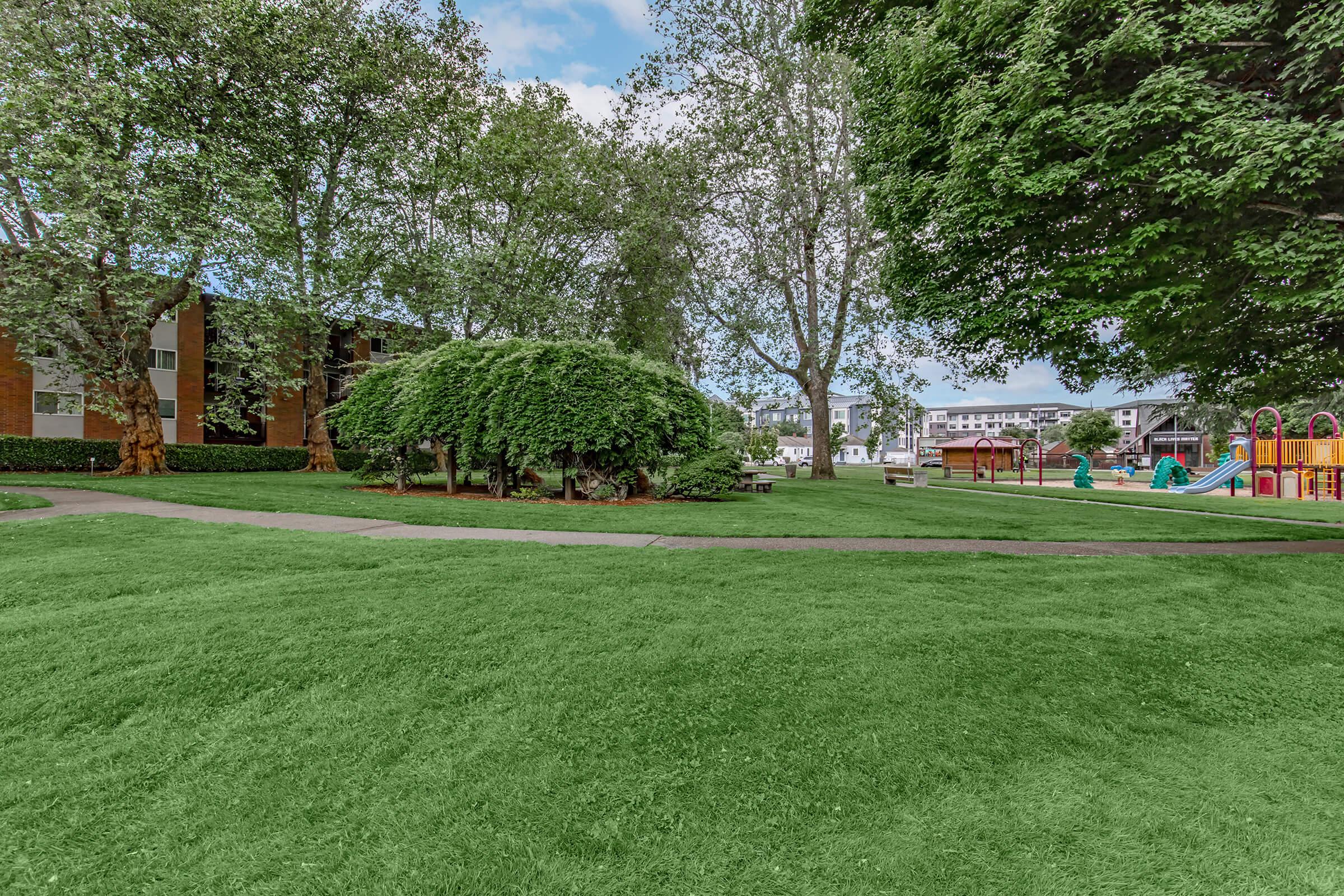 Lush green park with neatly trimmed grass, surrounded by trees. A pathway runs through the park, leading to a playground with colorful equipment in the background. Nearby buildings provide a residential backdrop to the tranquil outdoor space. The sky is clear with a few clouds.