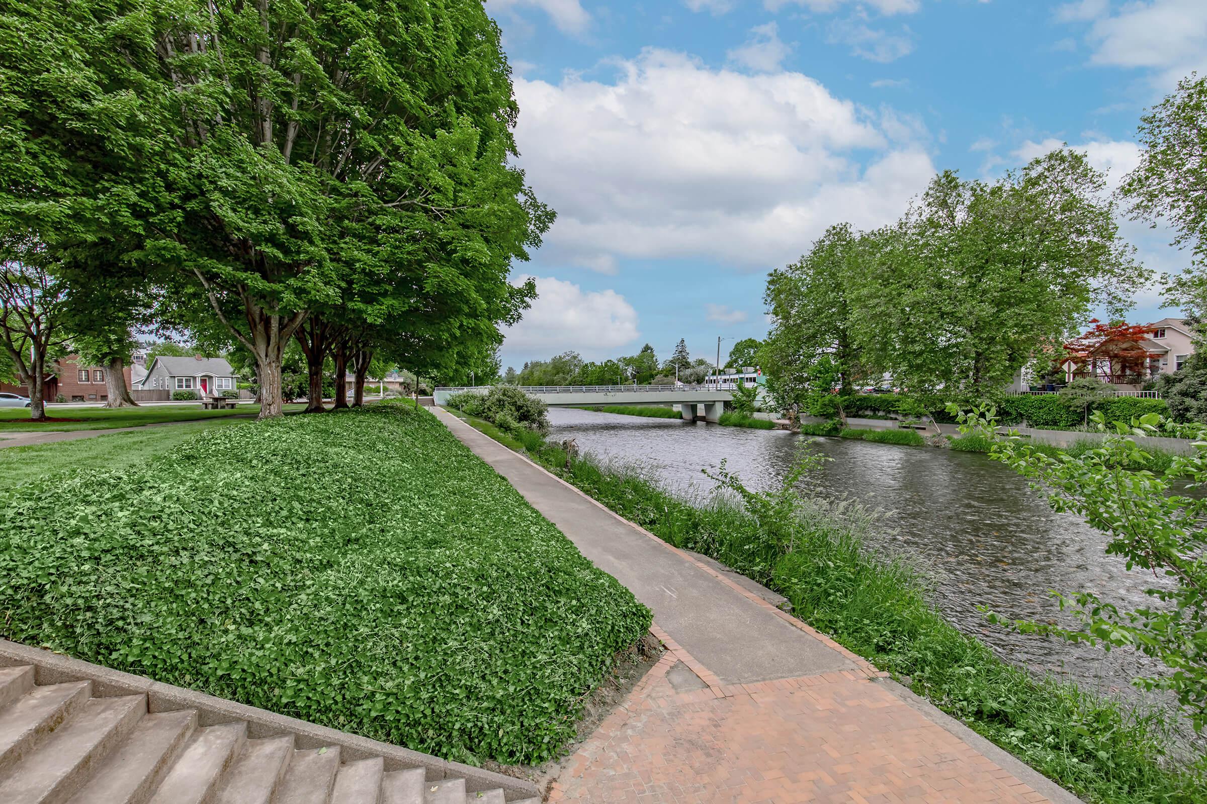 A serene riverside path lined with lush greenery, featuring trees on the left and a calm waterway on the right. A bridge is visible in the distance, connecting both sides of the river. The sky is partly cloudy, creating a peaceful outdoor scene perfect for leisurely walks.