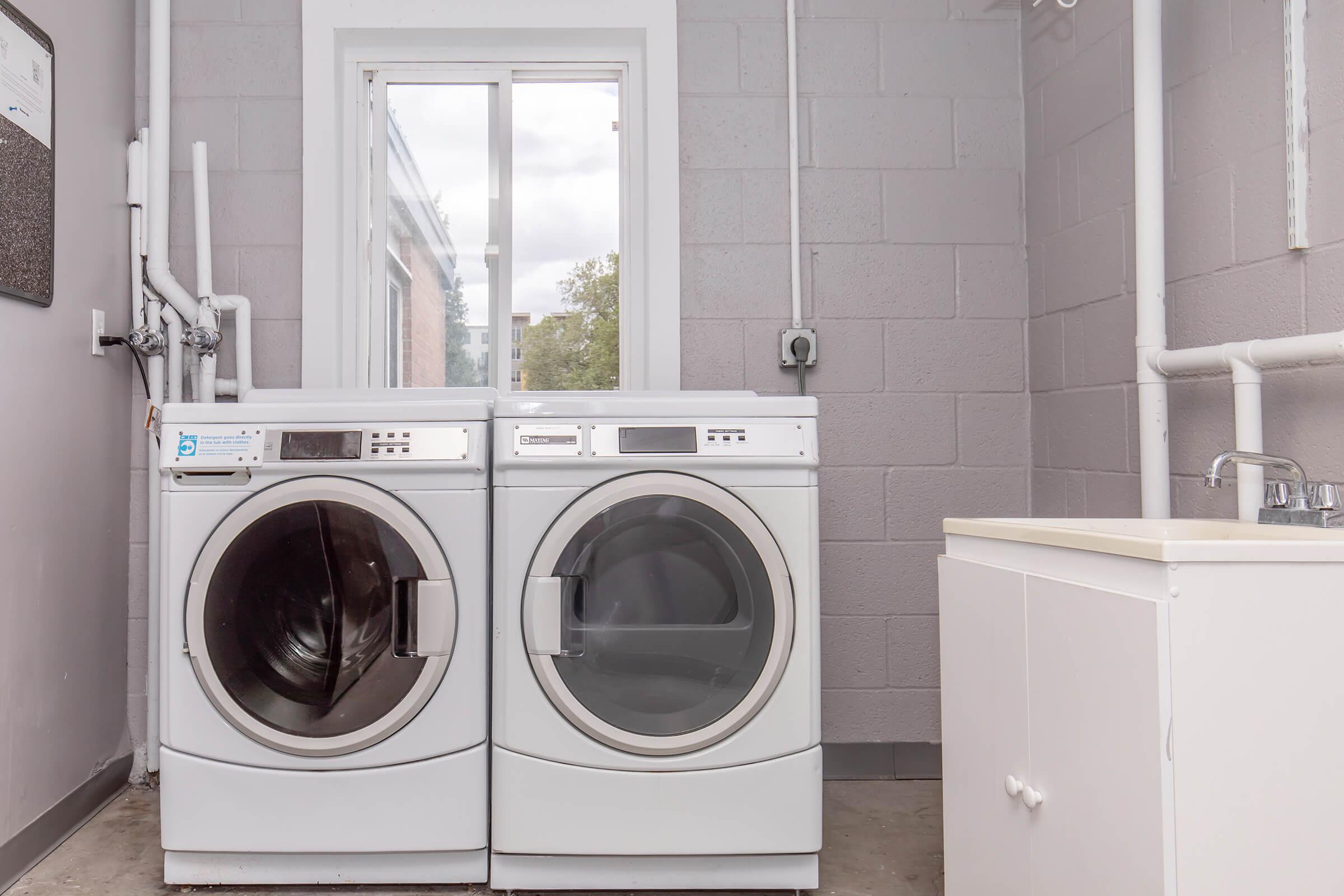 Two modern front-loading washing machines side by side in a well-lit laundry room. A window behind them allows natural light in, and a white countertop with a sink is visible to the right. The walls are gray, and pipes are exposed, adding a functional aesthetic to the space.