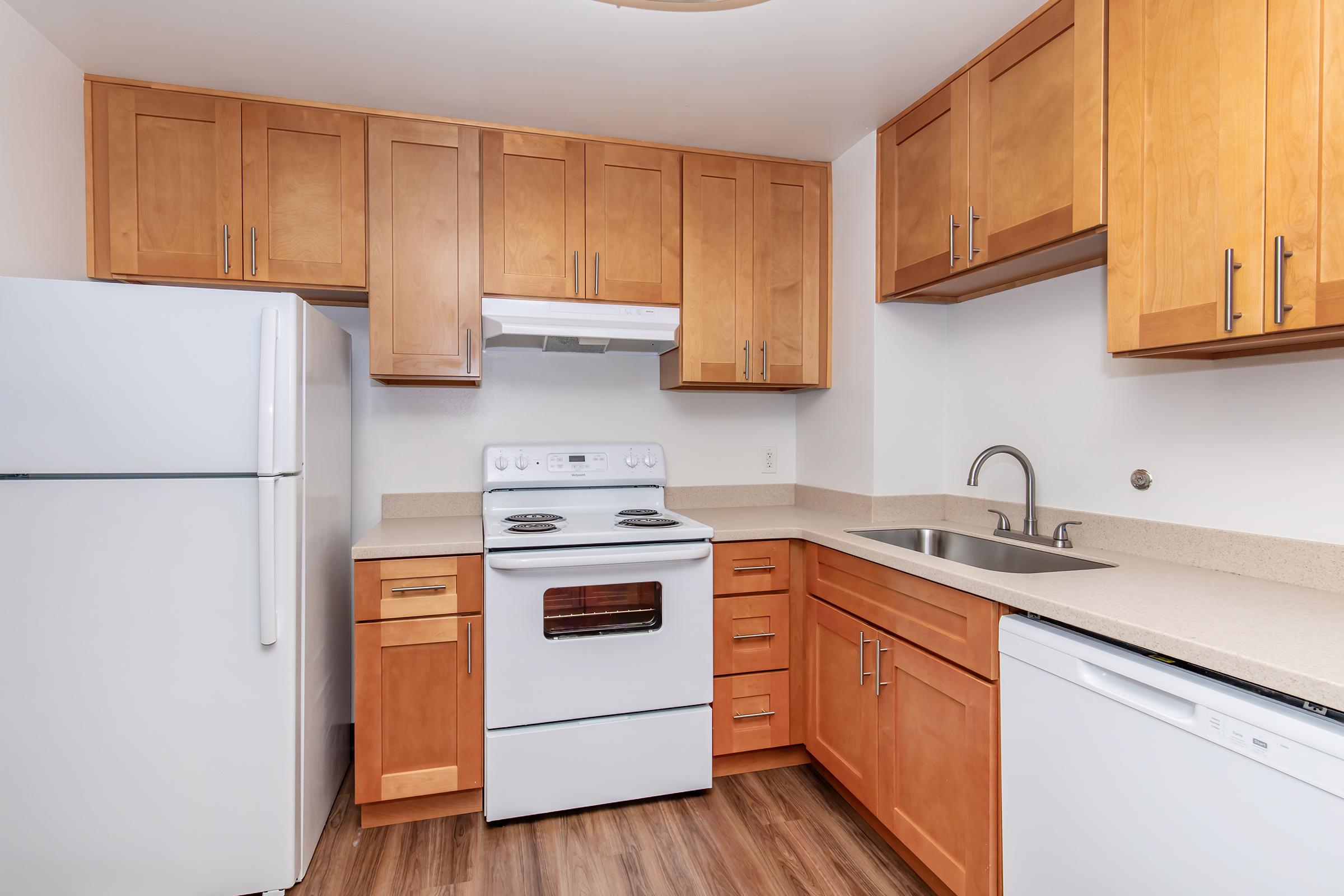 A modern kitchen featuring light wood cabinets, a white refrigerator, a white stove with an oven, a sink with a faucet, and a dishwasher. The kitchen has a clean, bright design with a neutral color scheme and wood-look flooring.