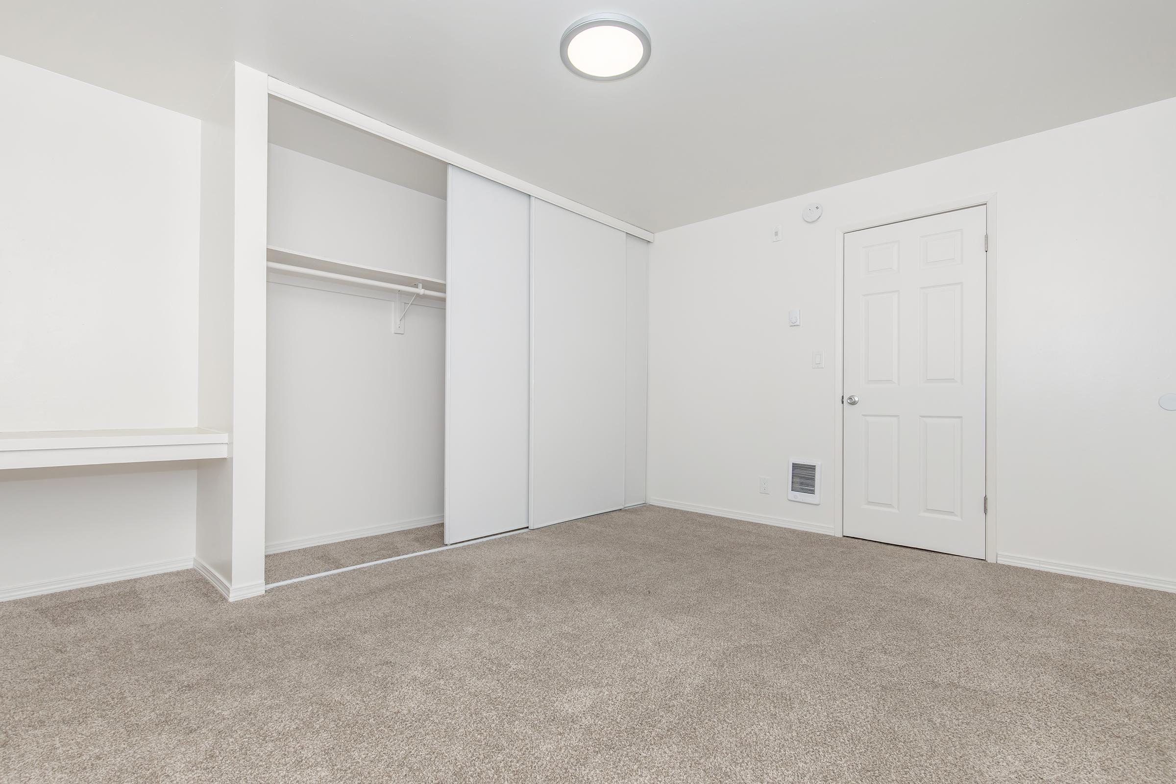 A spacious, empty room with light beige carpet, featuring a white closet with sliding doors, a plain white wall, and a door on the right. The room is well-lit by a ceiling light, and there is a small area with no furniture. The overall color scheme is neutral and clean, creating an open atmosphere.