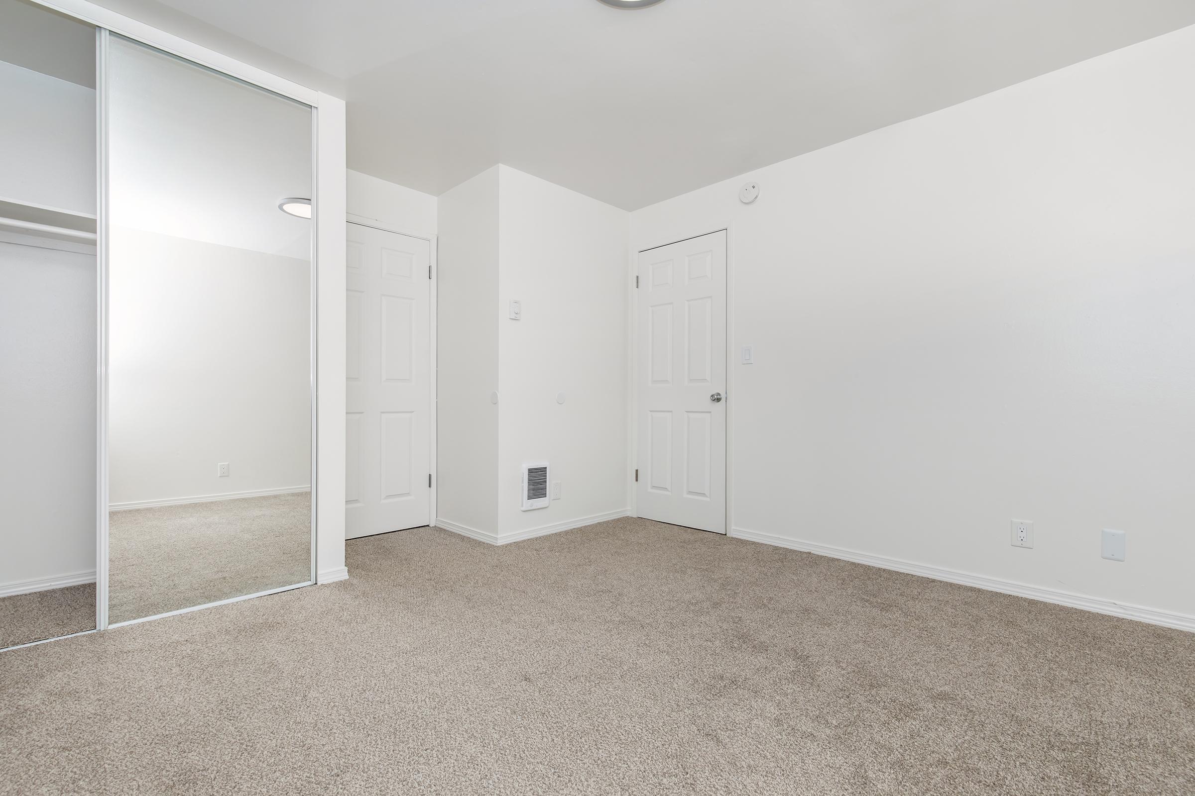 Empty, well-lit room with light beige carpet, white walls, and a full-length mirror on the left. There are two doors: one leading to a closet and the other an entrance, both white. The room features a ceiling light and a vent on the floor. Overall, the space appears clean and minimalistic.