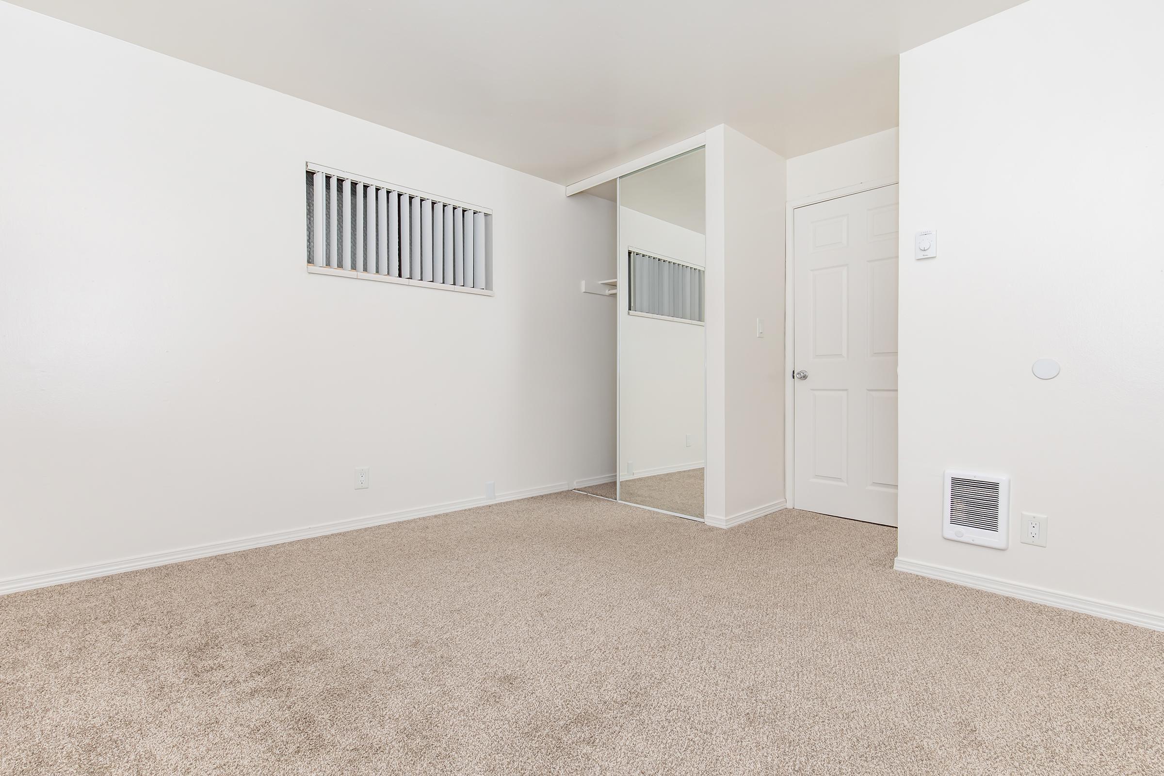 A spacious room with beige carpet flooring and white walls. It features a small, rectangular window with vertical blinds, a mirrored closet door, and a door leading to another area. The room is well-lit and empty, creating a fresh and neutral atmosphere.