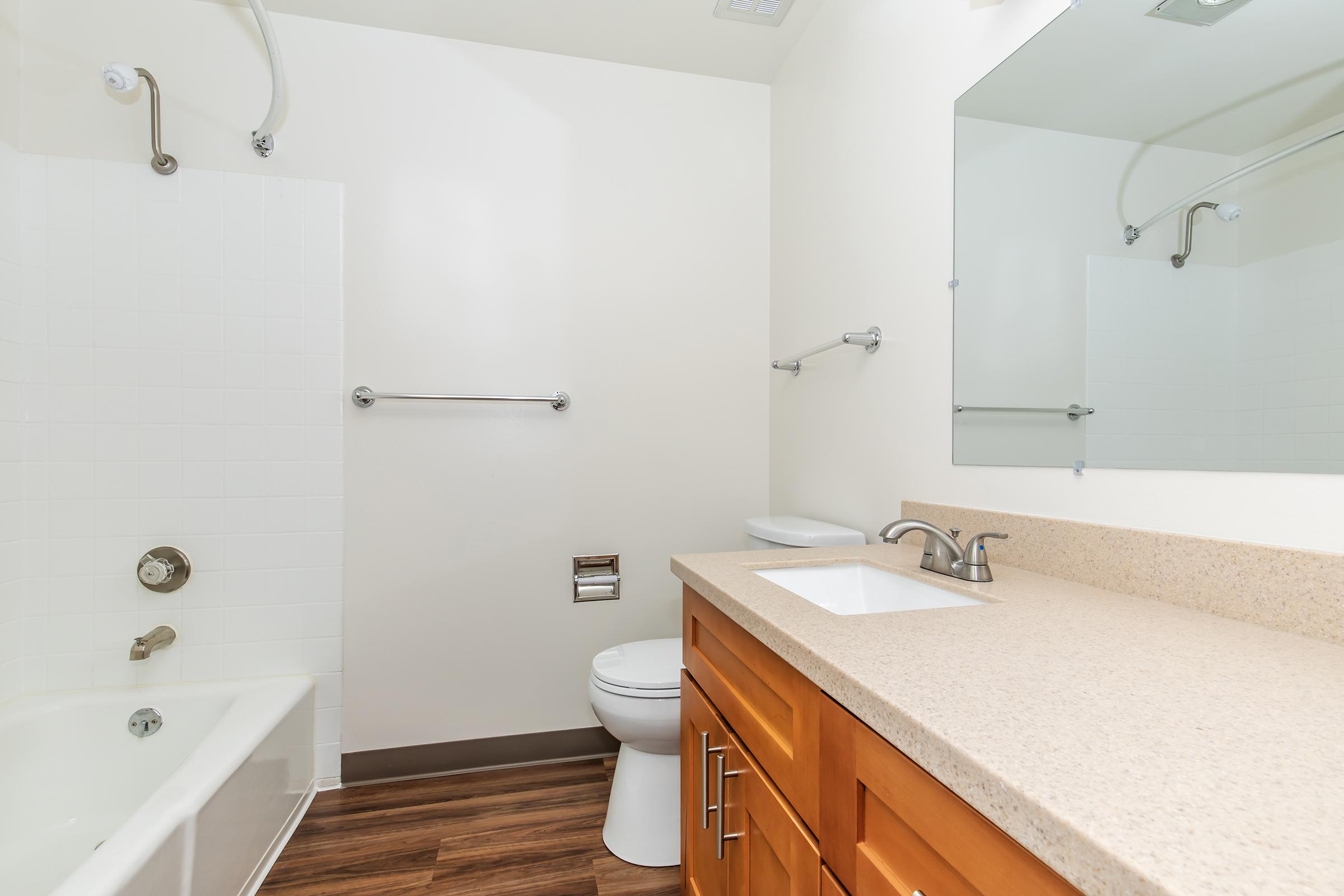 A modern bathroom featuring a bathtub with a shower, a wall-mounted toilet, and a single sink vanity with wooden cabinetry. The walls are painted light, and the floor is covered with laminate wood. A large mirror above the sink reflects the room's clean and minimalist design.