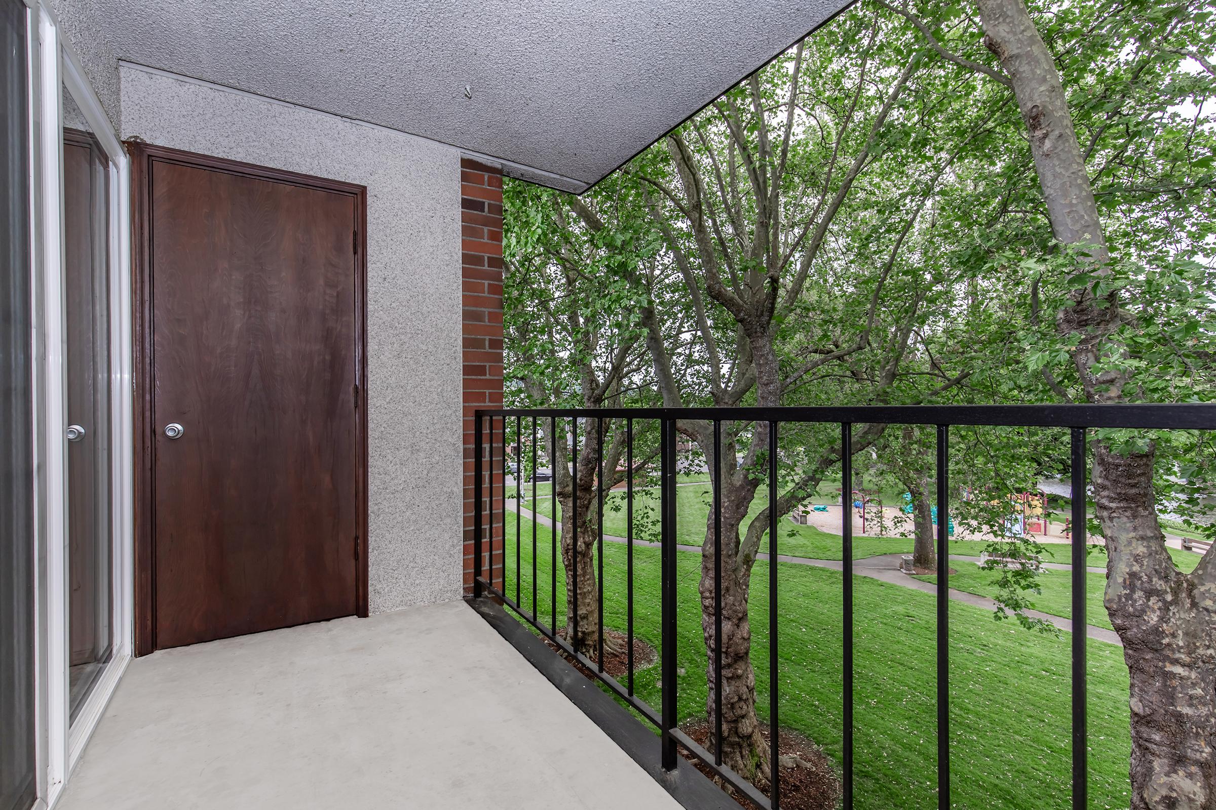 A small balcony featuring a black railing, with a view of green trees and a grassy area below. The balcony has a door leading inside, and a brown wooden door adjacent to it. The space appears clean and well-maintained, ideal for relaxing outdoors.