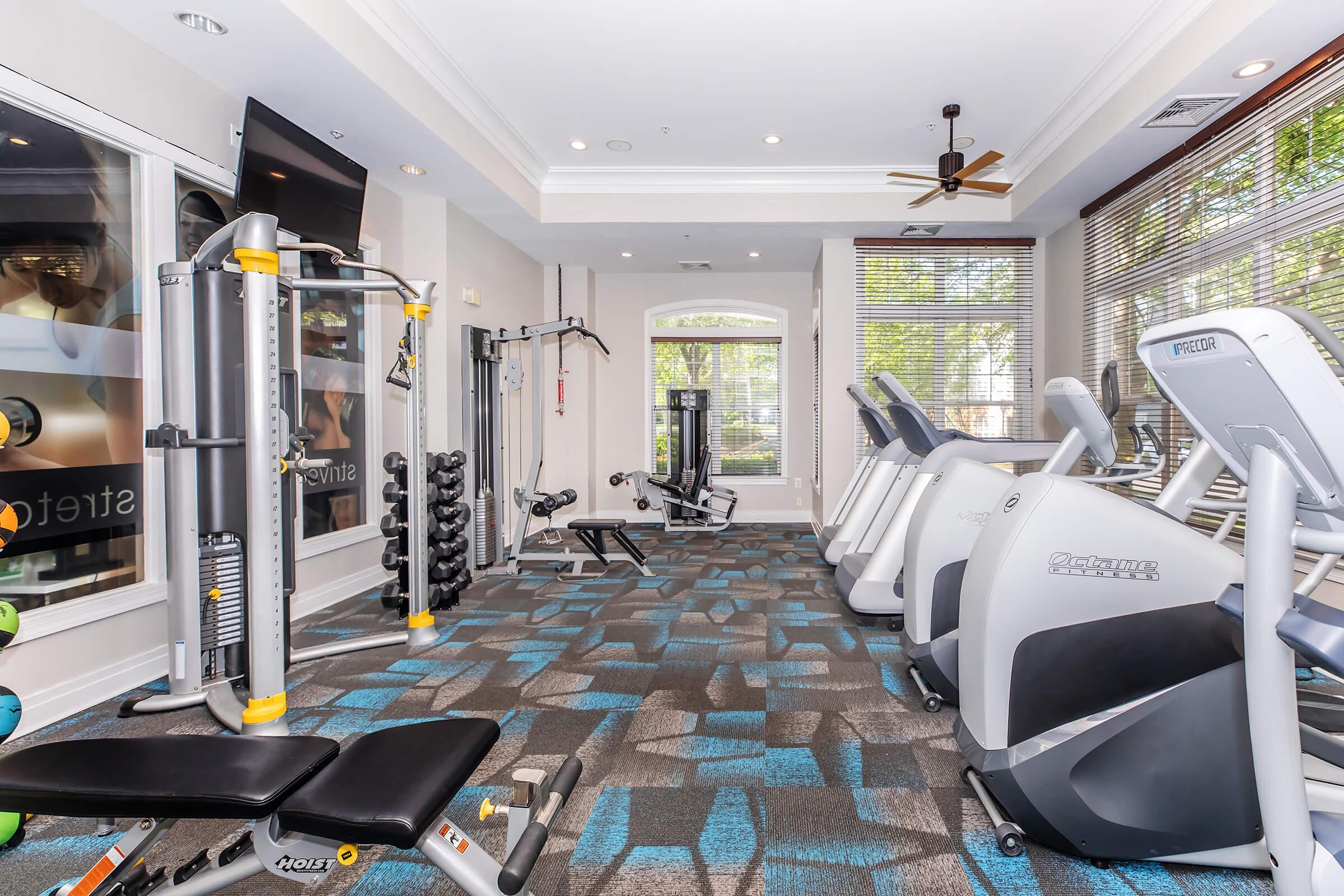 A modern fitness center featuring various exercise equipment, including stationary bikes, weight machines, and free weights. The gym is brightly lit with large windows and a cozy atmosphere, complemented by a ceiling fan and stylish carpeting.