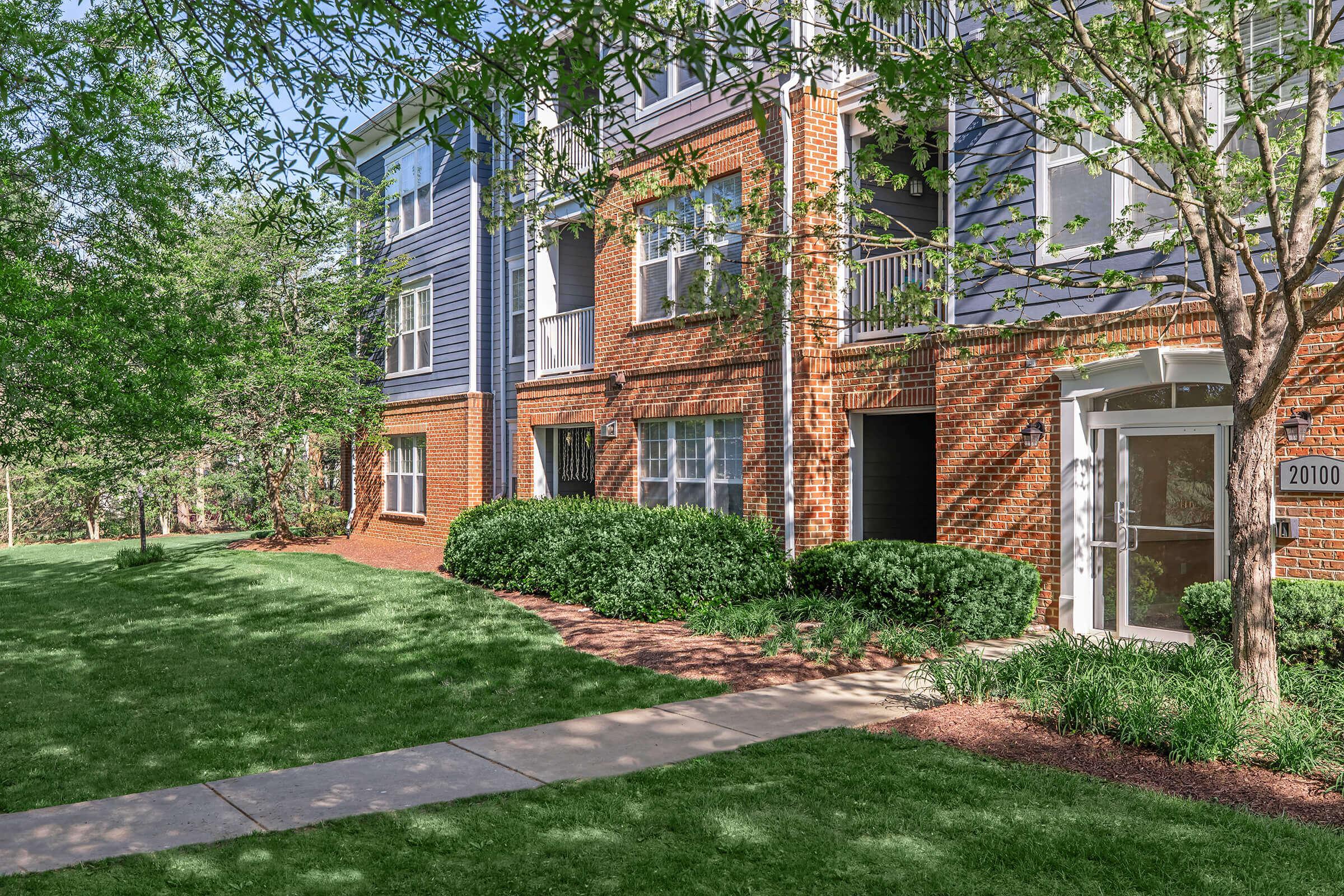 A well-maintained residential building with a mix of blue and red brick exterior. The ground features a manicured lawn with bushes and trees. A paved walkway leads to the entrance, emphasizing the inviting atmosphere of the property. The setting is bright and surrounded by greenery.