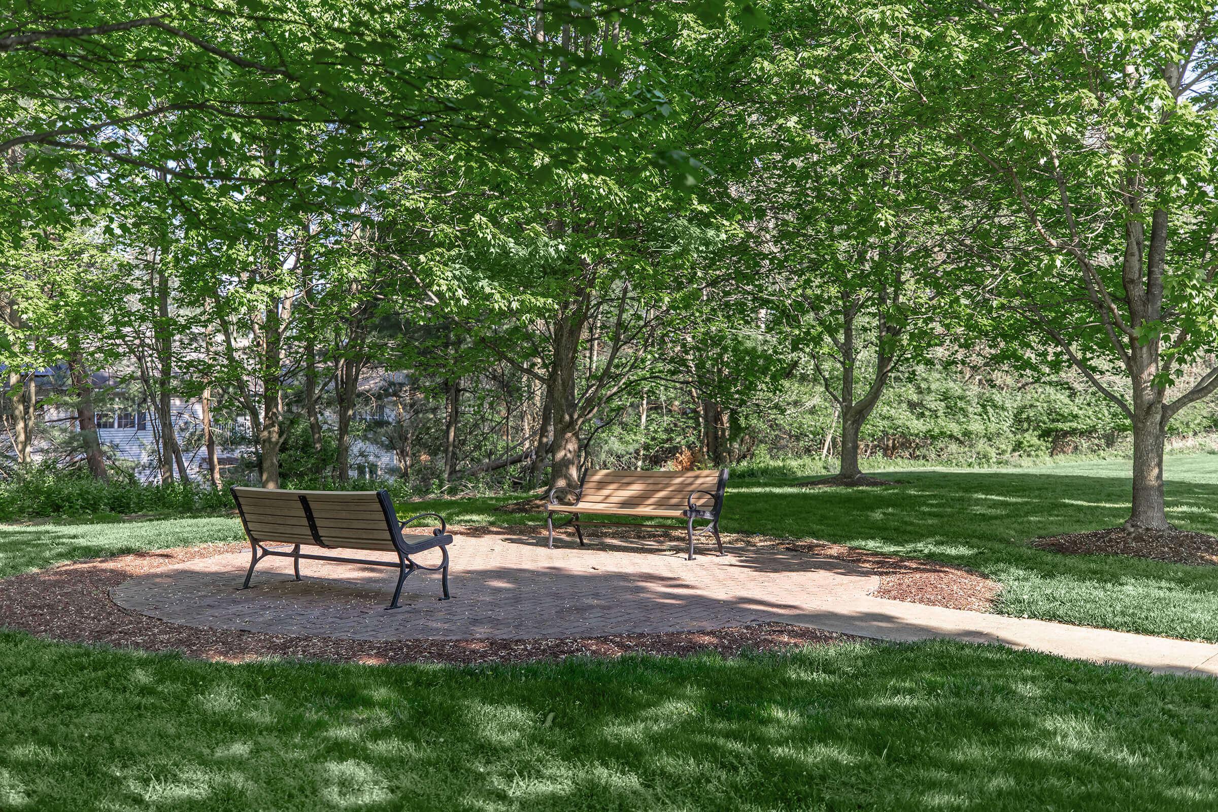 A serene park scene featuring two benches situated on a circular stone path surrounded by lush greenery and trees. The grassy area invites visitors to enjoy the peaceful outdoor space.