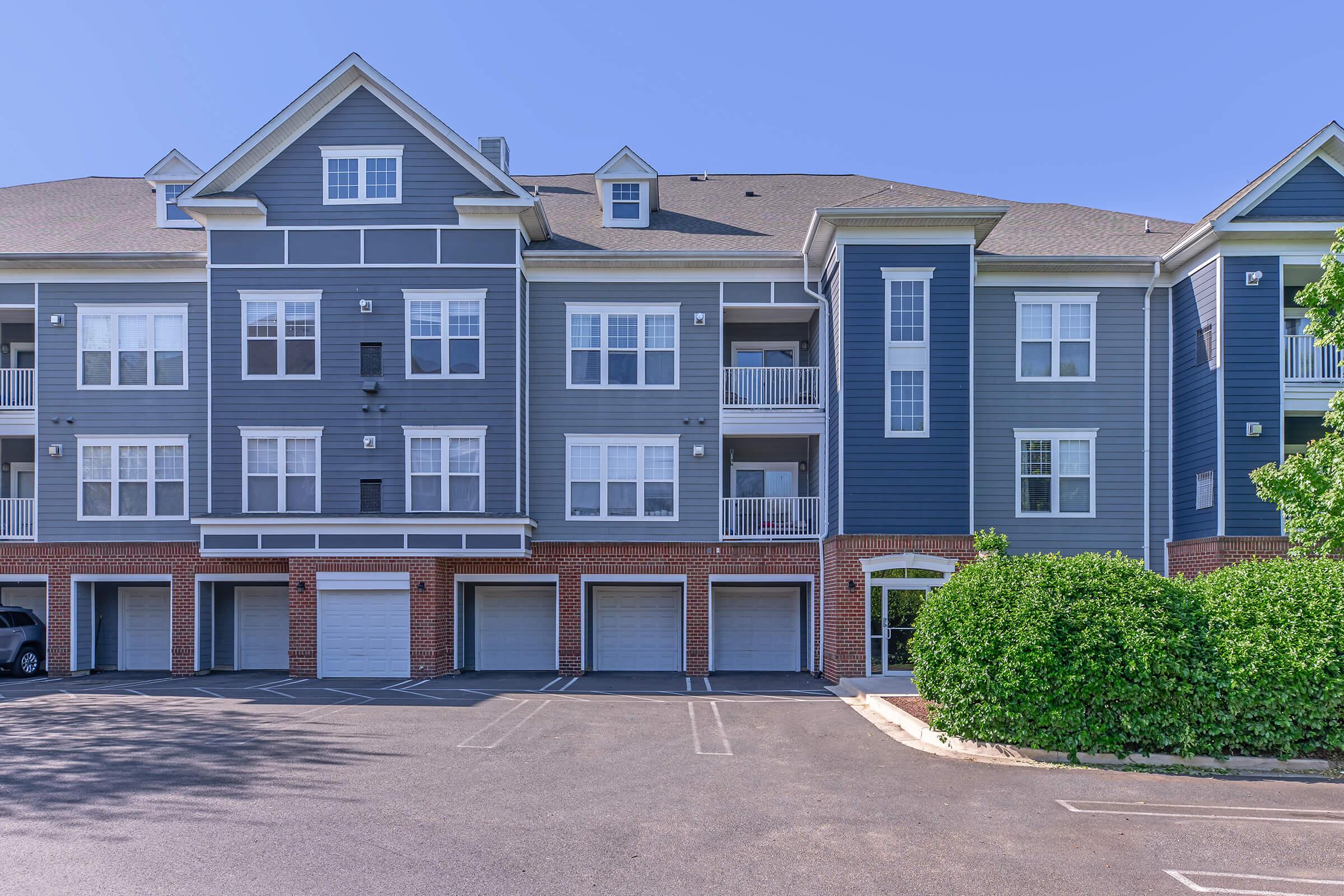 A modern apartment complex featuring a blue exterior with white trim, multiple balconies, and garage spaces on the ground level. The parking area is visible in front, with a well-maintained landscape including greenery. The sky is clear and blue.