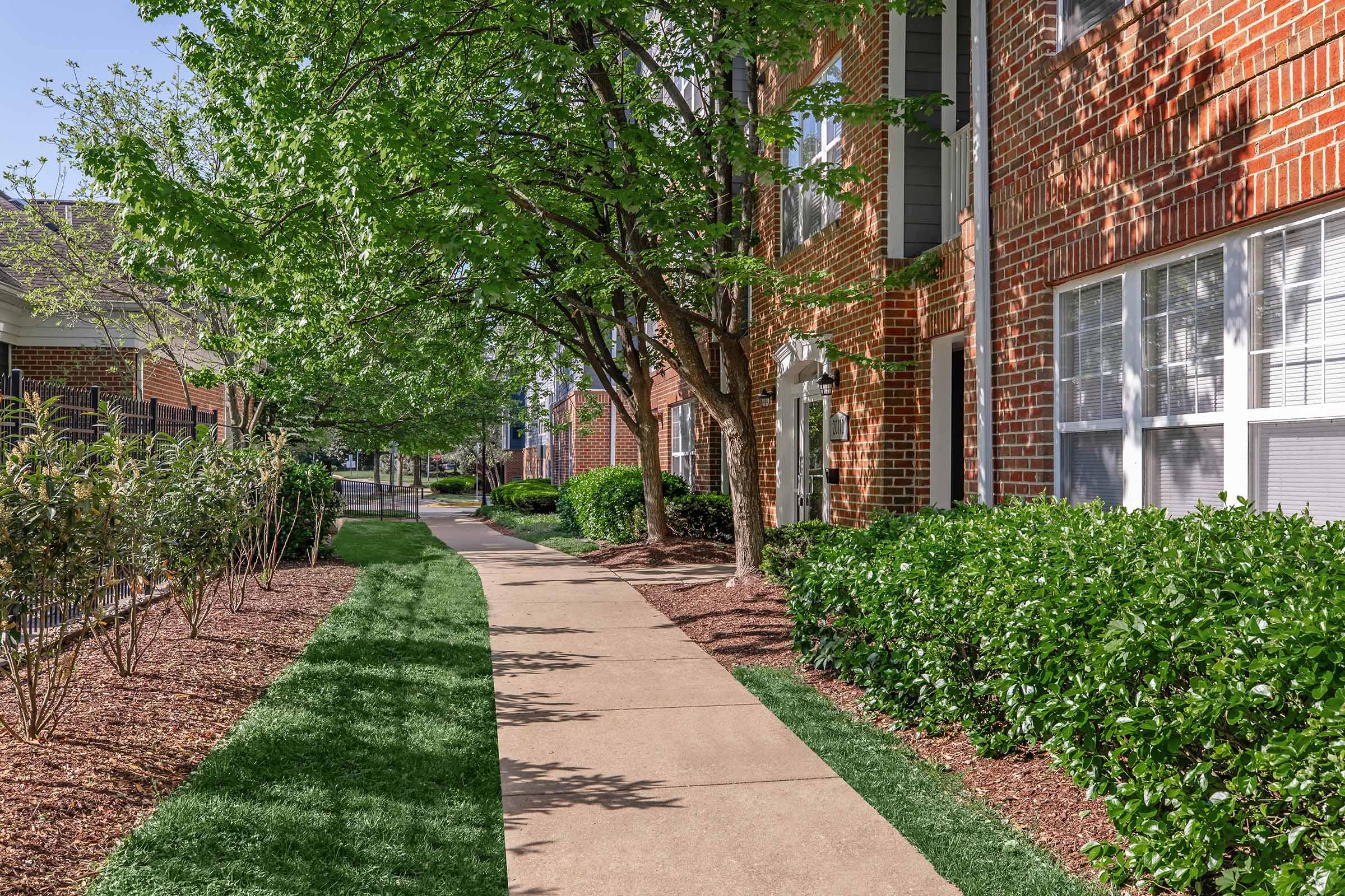 A well-maintained walkway lined with neatly trimmed bushes and trees. The path leads alongside red brick apartment buildings with bright windows, surrounded by green grass and landscaping. Sunlight filters through the leaves, creating a pleasant outdoor atmosphere.