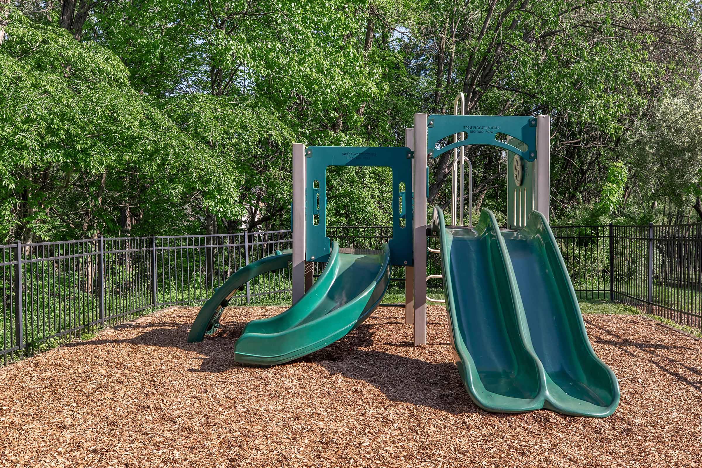 Two green slides on a playground structure surrounded by a wooden mulch surface and a fenced area, with lush green trees in the background. The playground set is designed for children, featuring a modern design and a safe play environment.