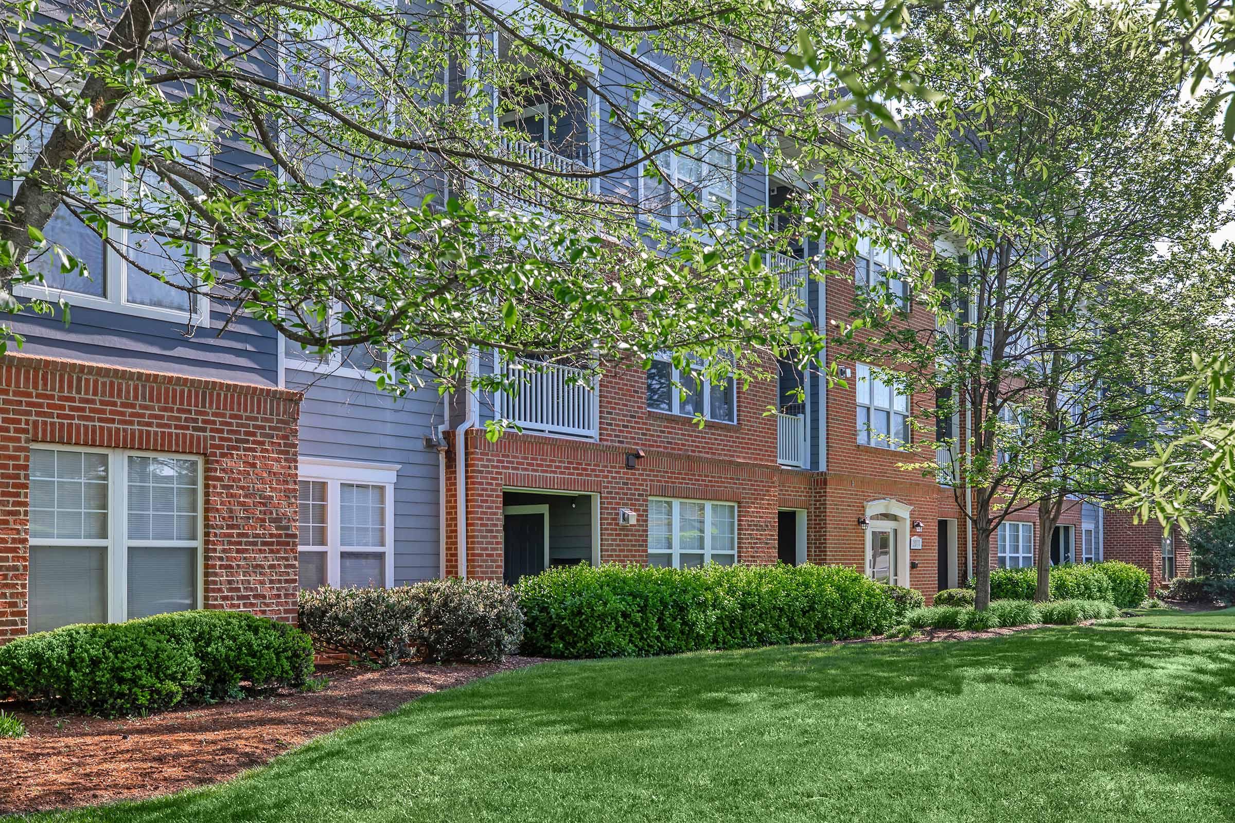 A well-maintained residential building with a mix of brick and blue siding. The facade features multiple windows and balconies, surrounded by lush green grass and neatly trimmed bushes. Several trees provide shade, creating a pleasant outdoor environment.