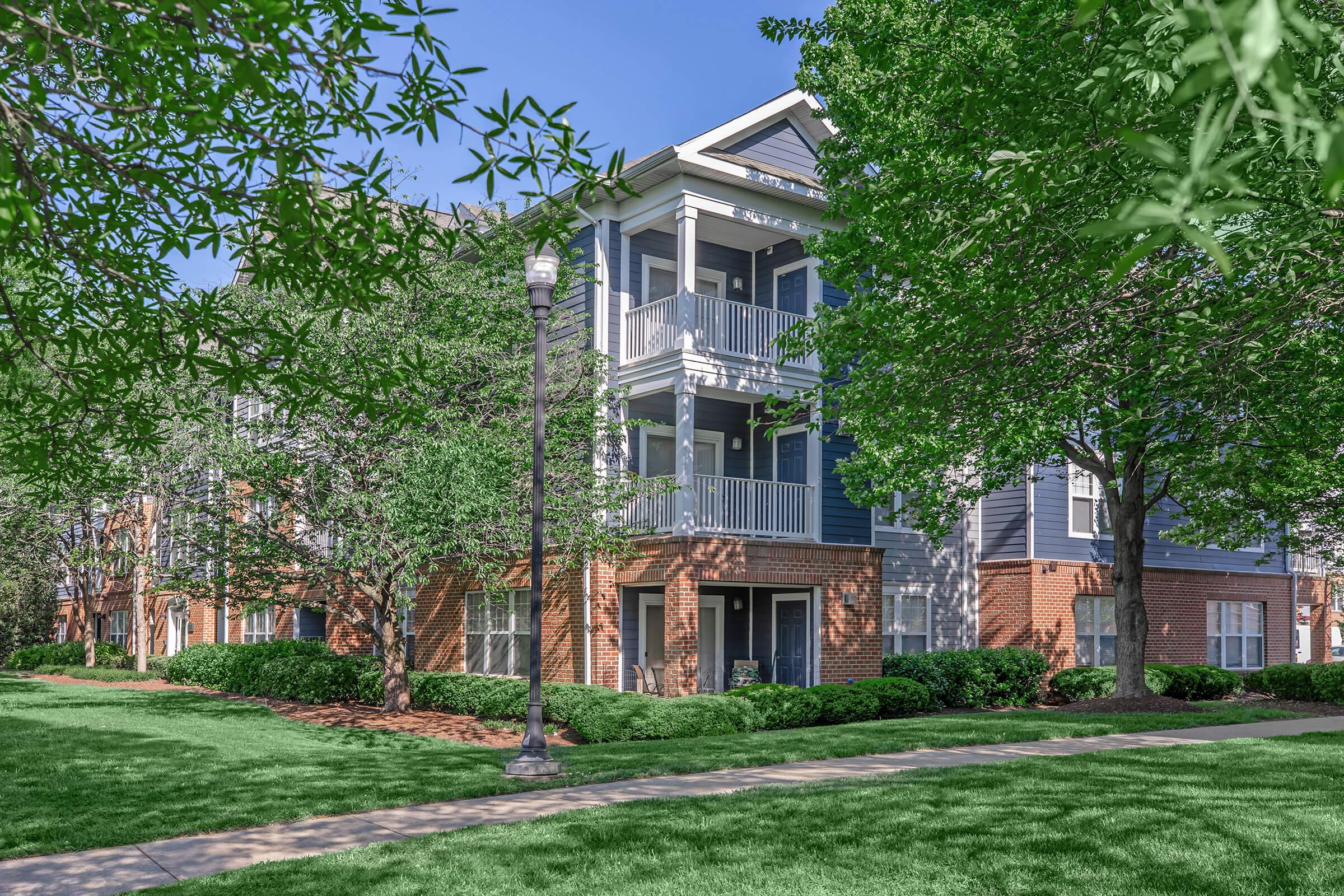 A three-story apartment building with a mix of brick and siding, featuring several balconies. The spacious grounds are well-maintained with lush green grass, small shrubs, and trees surrounding the building, creating a welcoming residential environment on a sunny day.