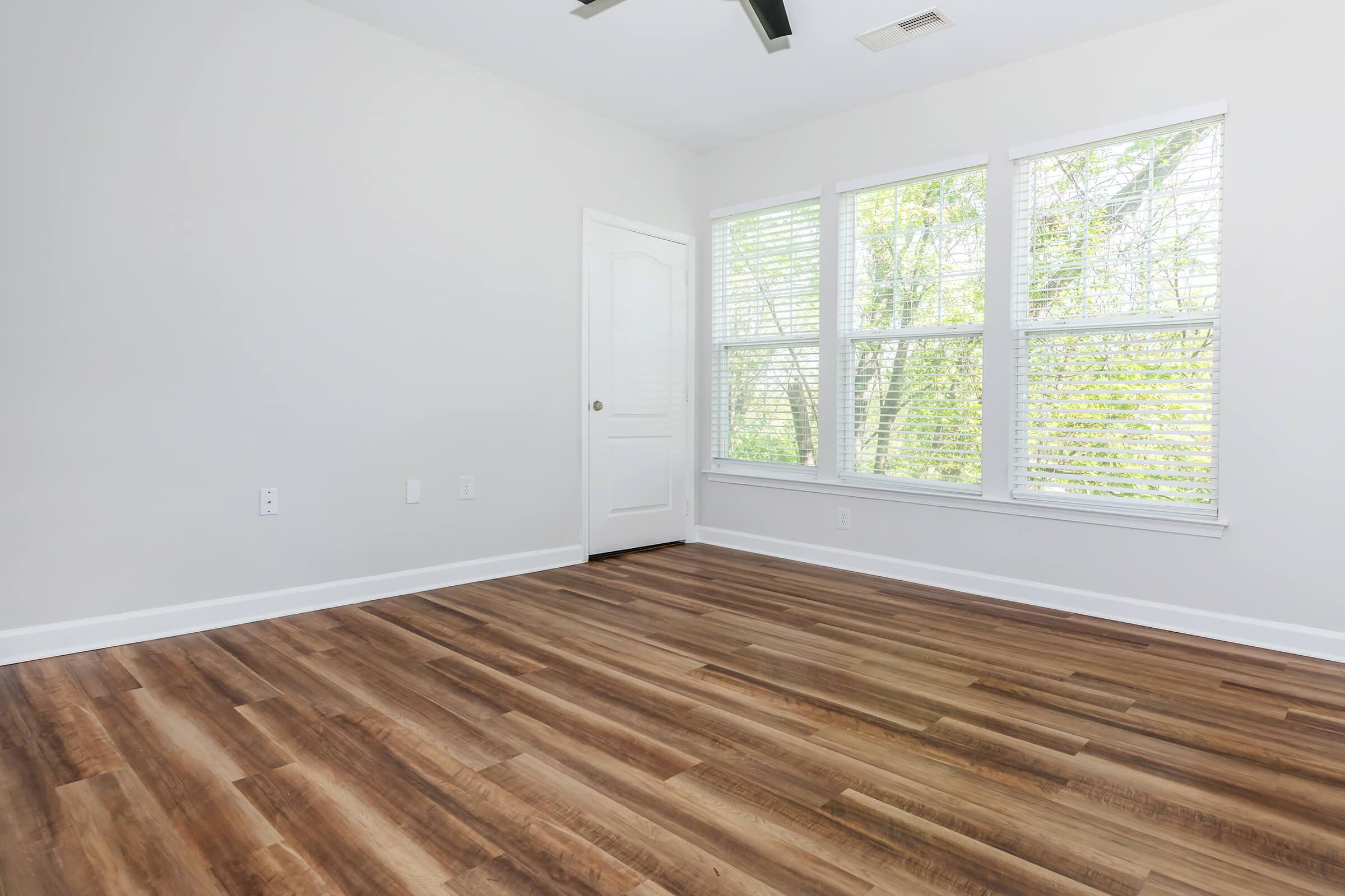 Empty room with large windows allowing natural light, featuring wooden flooring and a light-colored wall. A door is visible on the side, and no furniture is present, creating a spacious and airy atmosphere.