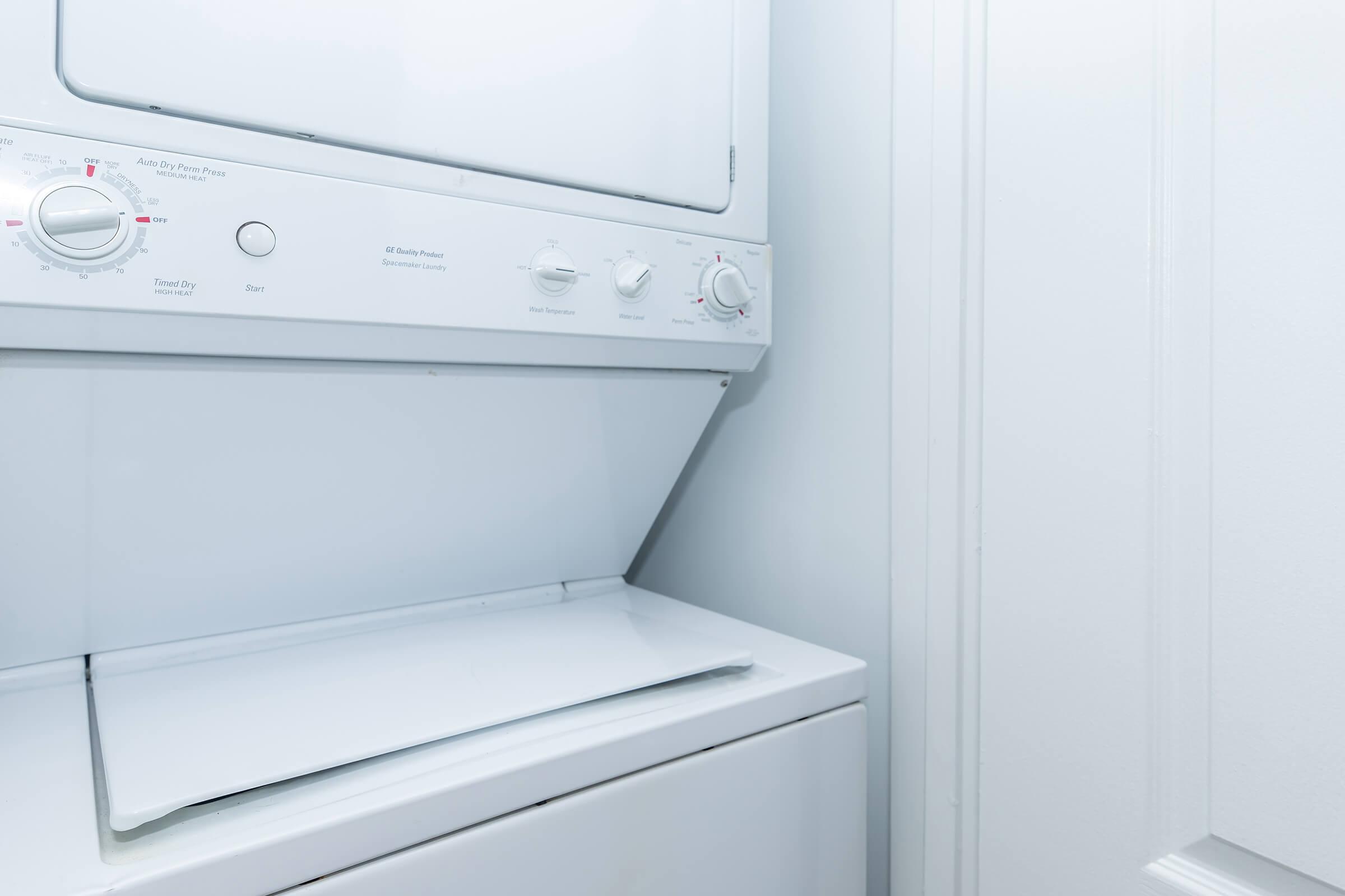 A stacked washer and dryer unit in a laundry room, featuring a white exterior and control knobs for various settings. The walls are plain and light-colored, creating a clean and minimalist appearance.