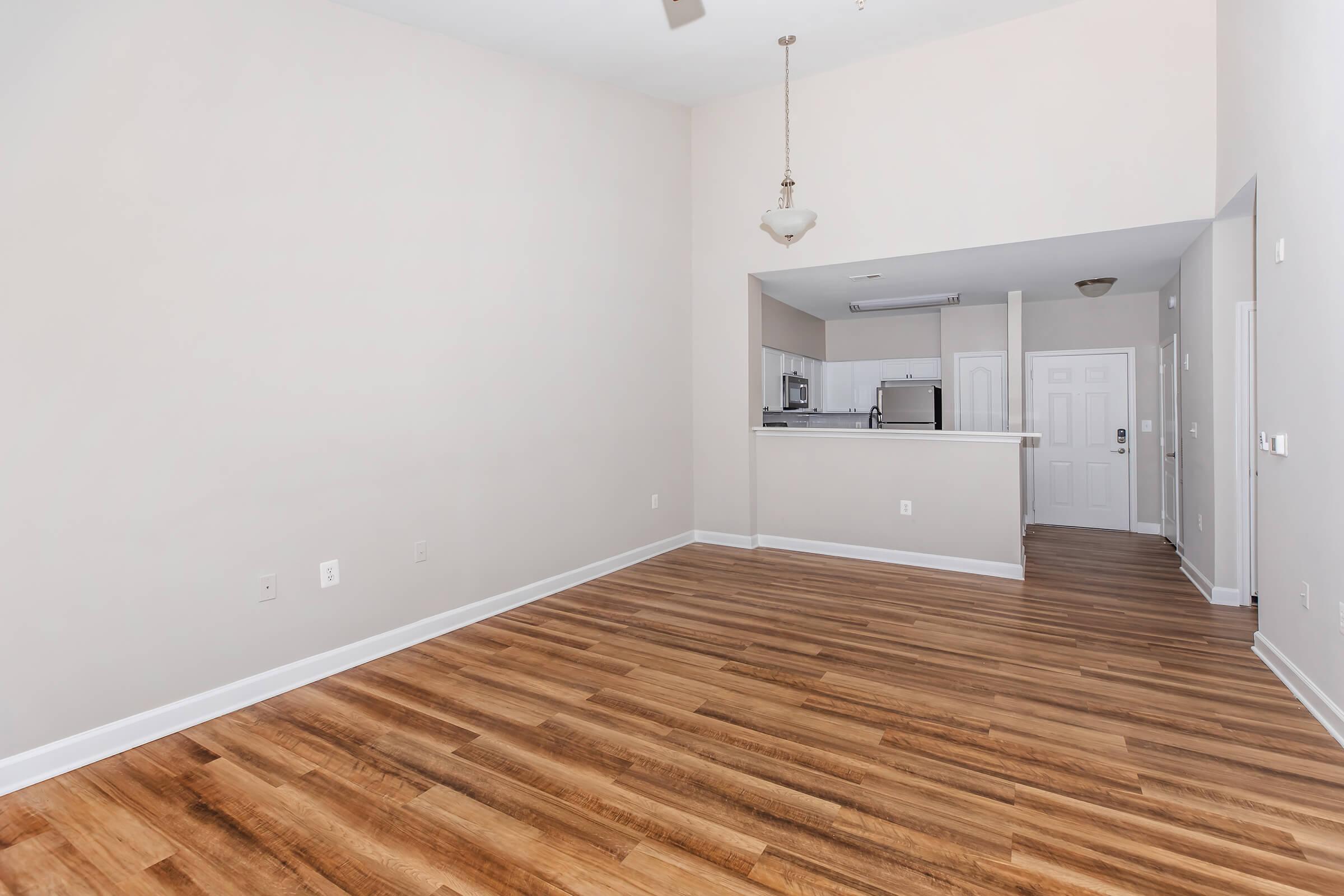 Spacious interior of an apartment with light-colored walls and wooden flooring. The area features a ceiling fan, a kitchen visible in the background, and a front door. Natural light fills the room, creating a bright and open atmosphere, ideal for decorating and personalizing.