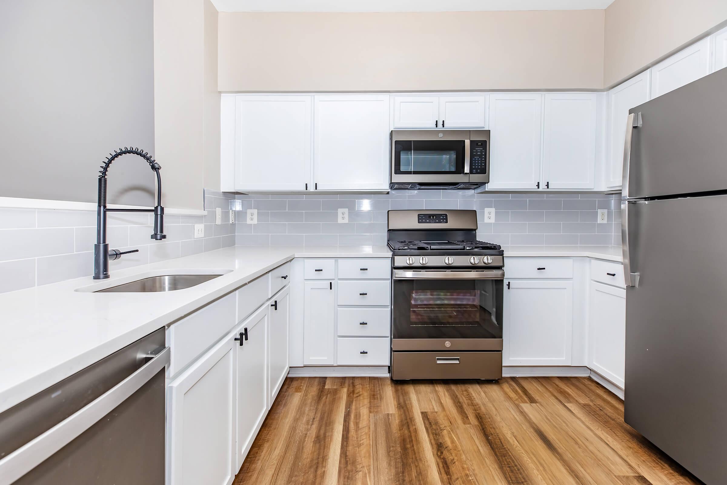 A modern kitchen featuring white cabinetry, a stainless steel refrigerator, and a gas stove with a microwave above. The countertop is a sleek white, and there's a single basin sink with a stylish faucet. The flooring is wood-like laminate, creating a warm and inviting atmosphere.