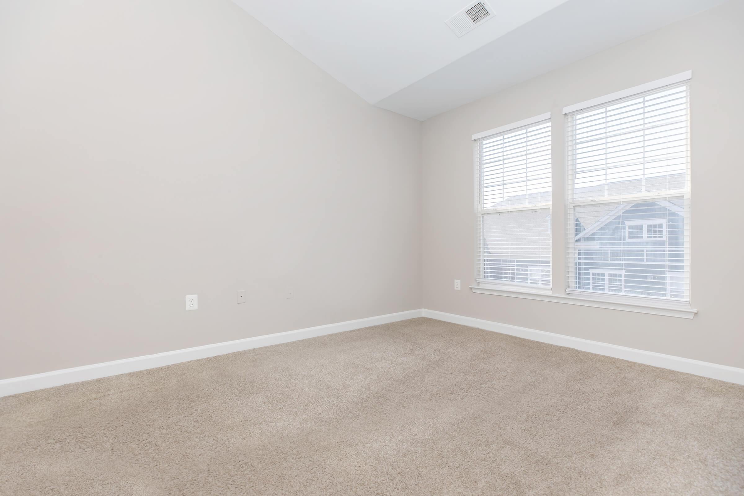 Empty interior room with beige walls and a light carpet. Two windows with white blinds allow natural light to enter. The corner reveals part of a blue building outside, creating a bright and airy atmosphere.