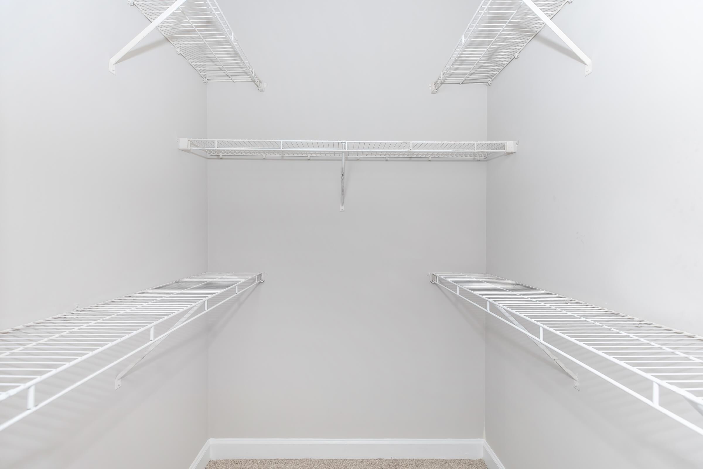 Empty walk-in closet featuring white wire shelving against light gray walls. The closet has multiple shelves, providing ample space for storage, and is carpeted. The overall look is clean and minimalist.