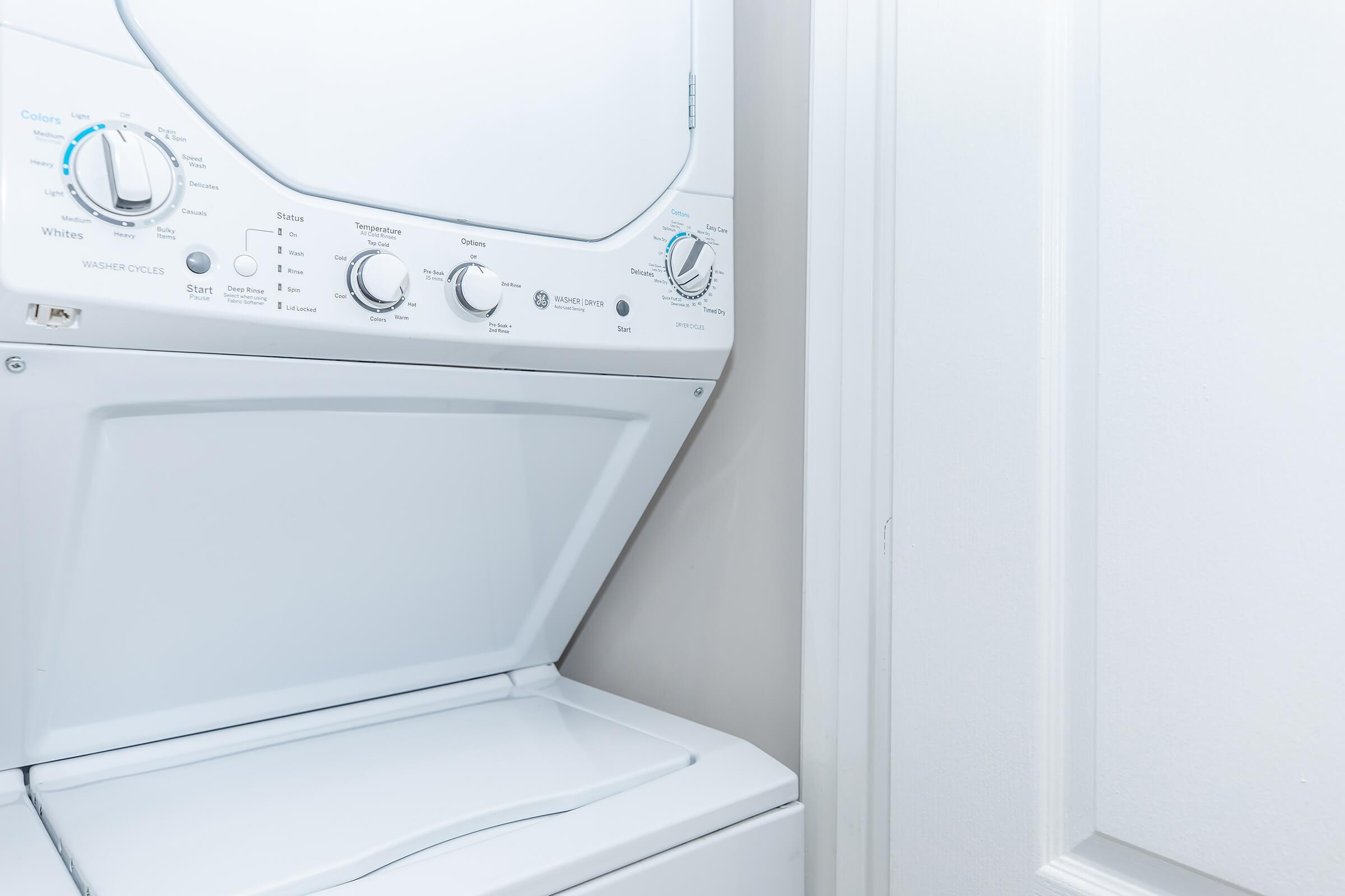 A stacked washer and dryer unit in a laundry room, with control knobs for different settings. The appliance is white and positioned next to a closed door. The overall setting is clean and modern.