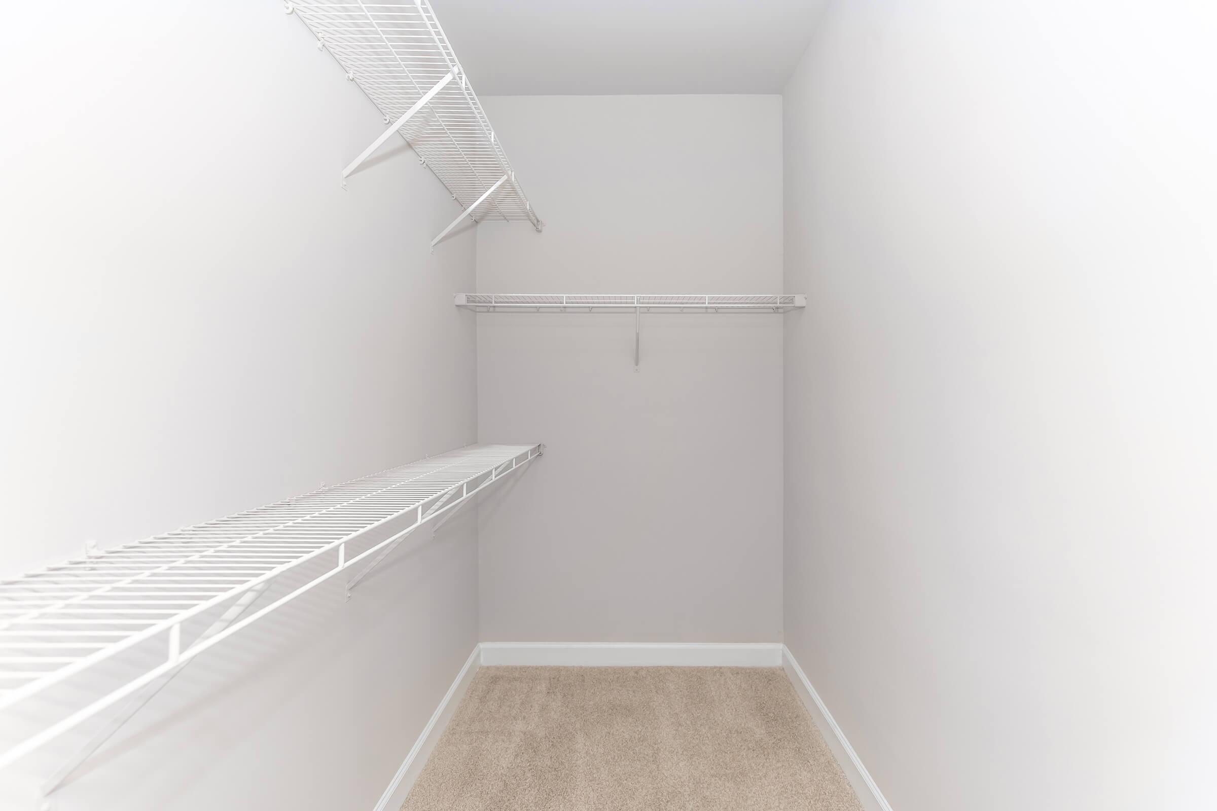Empty walk-in closet with light gray walls and beige carpet. Features two wire shelving units on the left and right walls, with ample space for storage. The area is well-lit and uncluttered, ideal for organizing clothes and accessories.