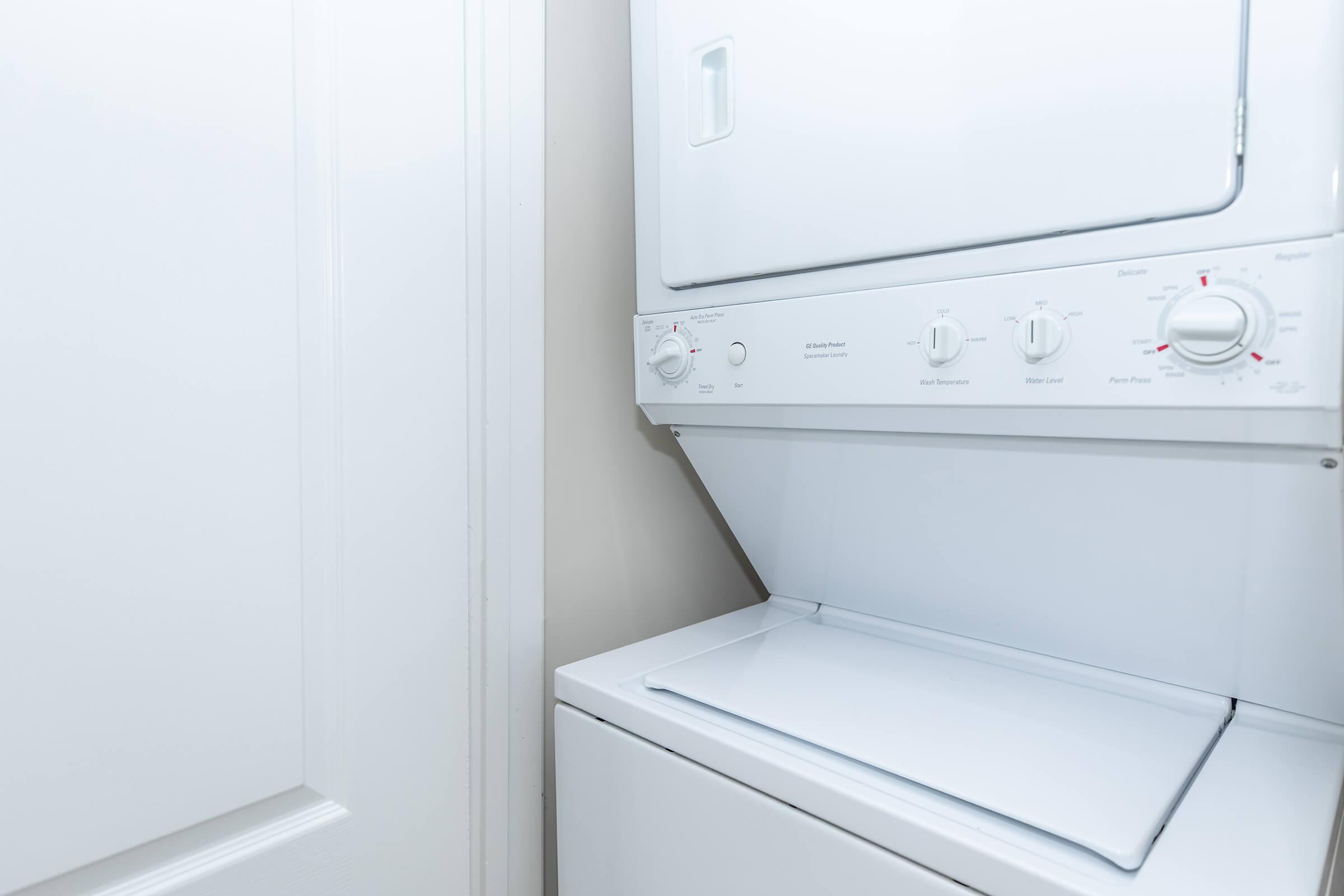 A stacked laundry unit featuring a washing machine on the bottom and a dryer on top, with control knobs and settings visible. The unit is positioned against a light-colored wall, with a door slightly visible nearby.