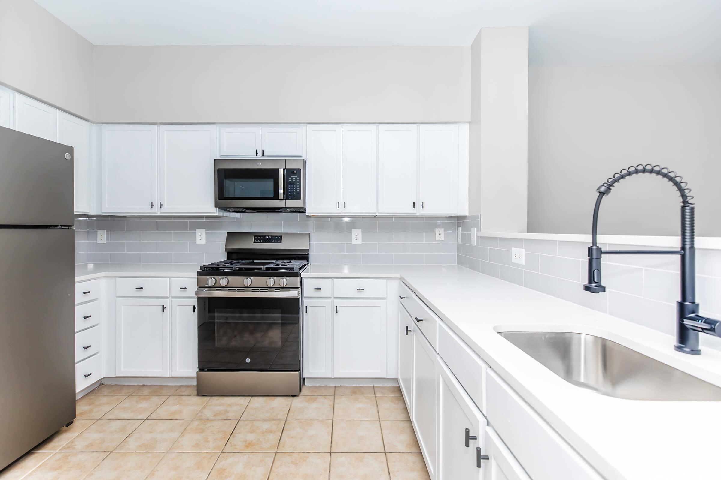 A modern kitchen featuring white cabinets, a stainless steel refrigerator, a black gas stove, and a microwave above it. The countertop is white, and there is a sleek black faucet above a stainless steel sink. The walls are light gray, and the floor is tiled with beige squares.