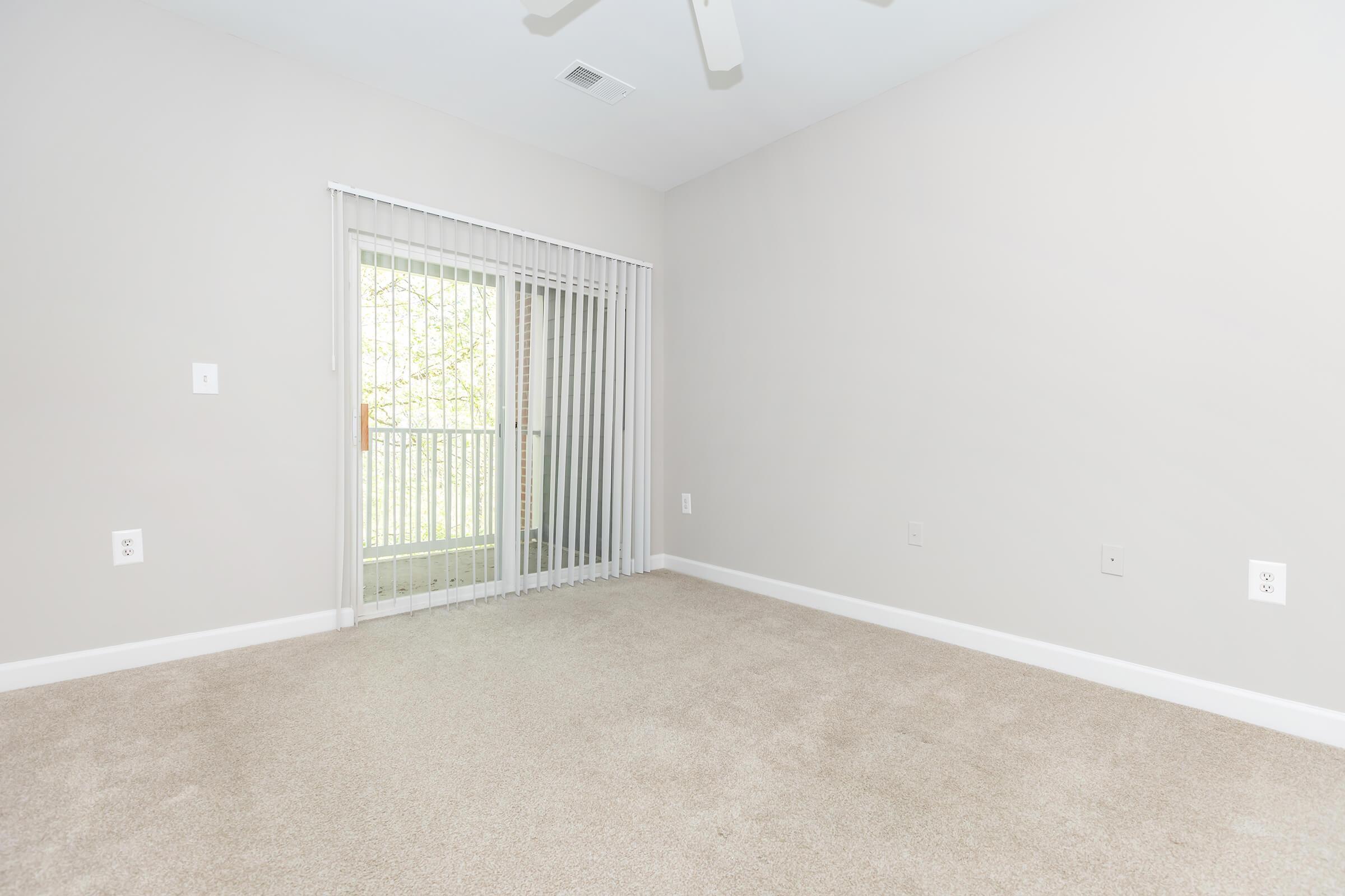 A spacious, empty room with light beige walls and plush carpet. A set of vertical blinds partially covers a sliding glass door leading to a balcony outside, allowing natural light to enter. The room has minimal decor and electrical outlets on the walls.