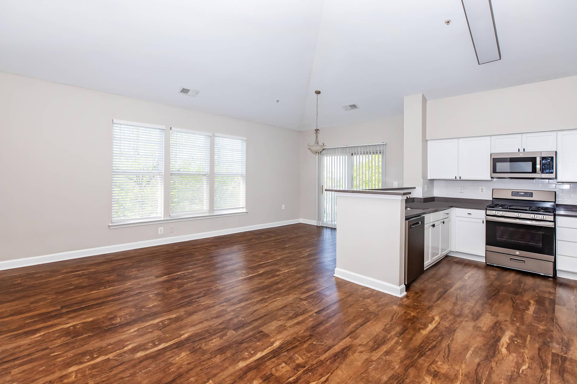 Spacious living area with hardwood-style flooring, large windows allowing natural light, and a modern kitchen featuring stainless steel appliances, a microwave, and a dining area. The open layout creates a bright and inviting atmosphere, perfect for entertaining or relaxing.