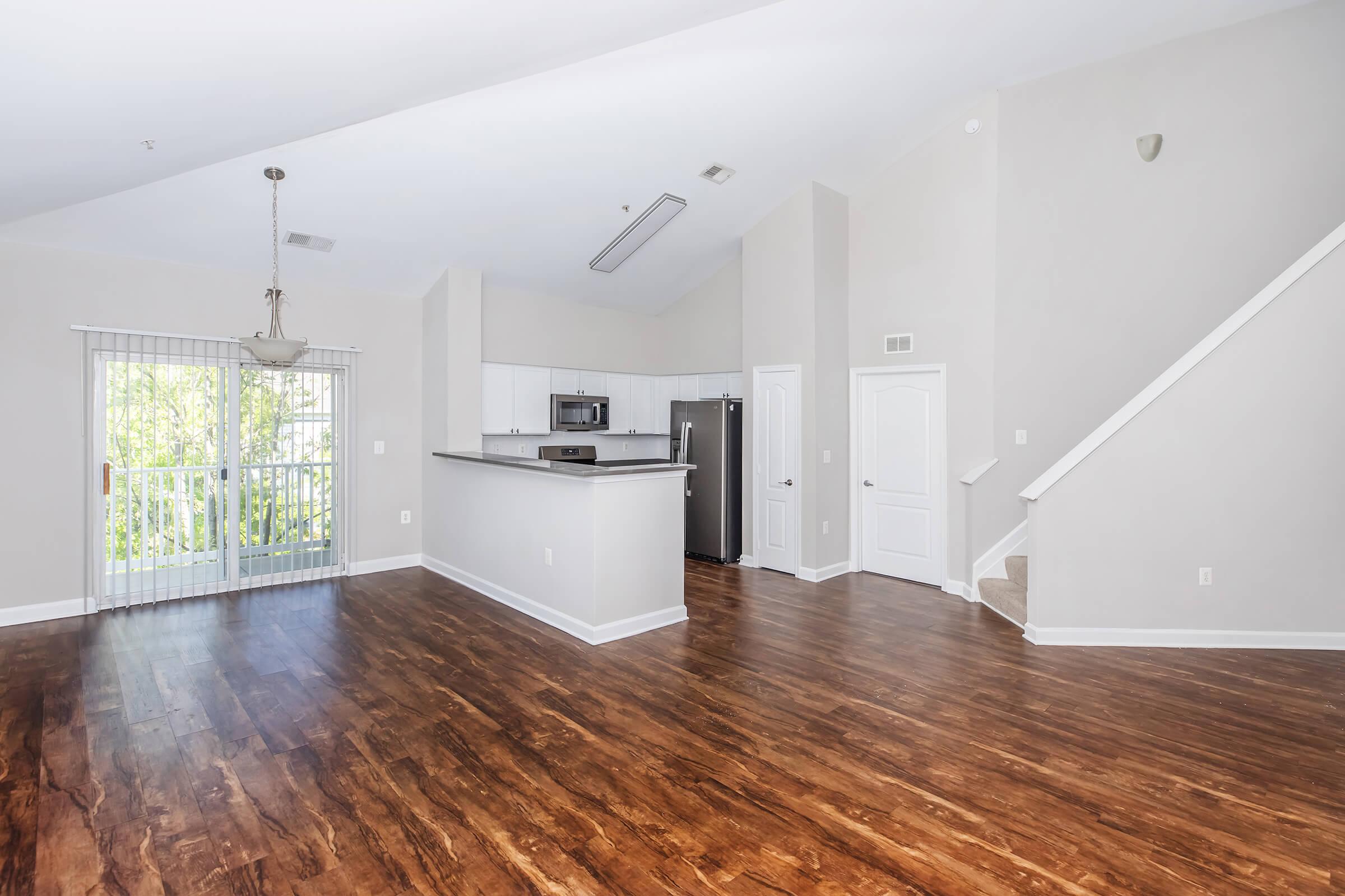 Spacious living area featuring hardwood floors, large windows with natural light, and a breakfast bar connecting to a modern kitchen with stainless steel appliances. The design includes a staircase leading to an upper level, and a sliding glass door opens to a small balcony.