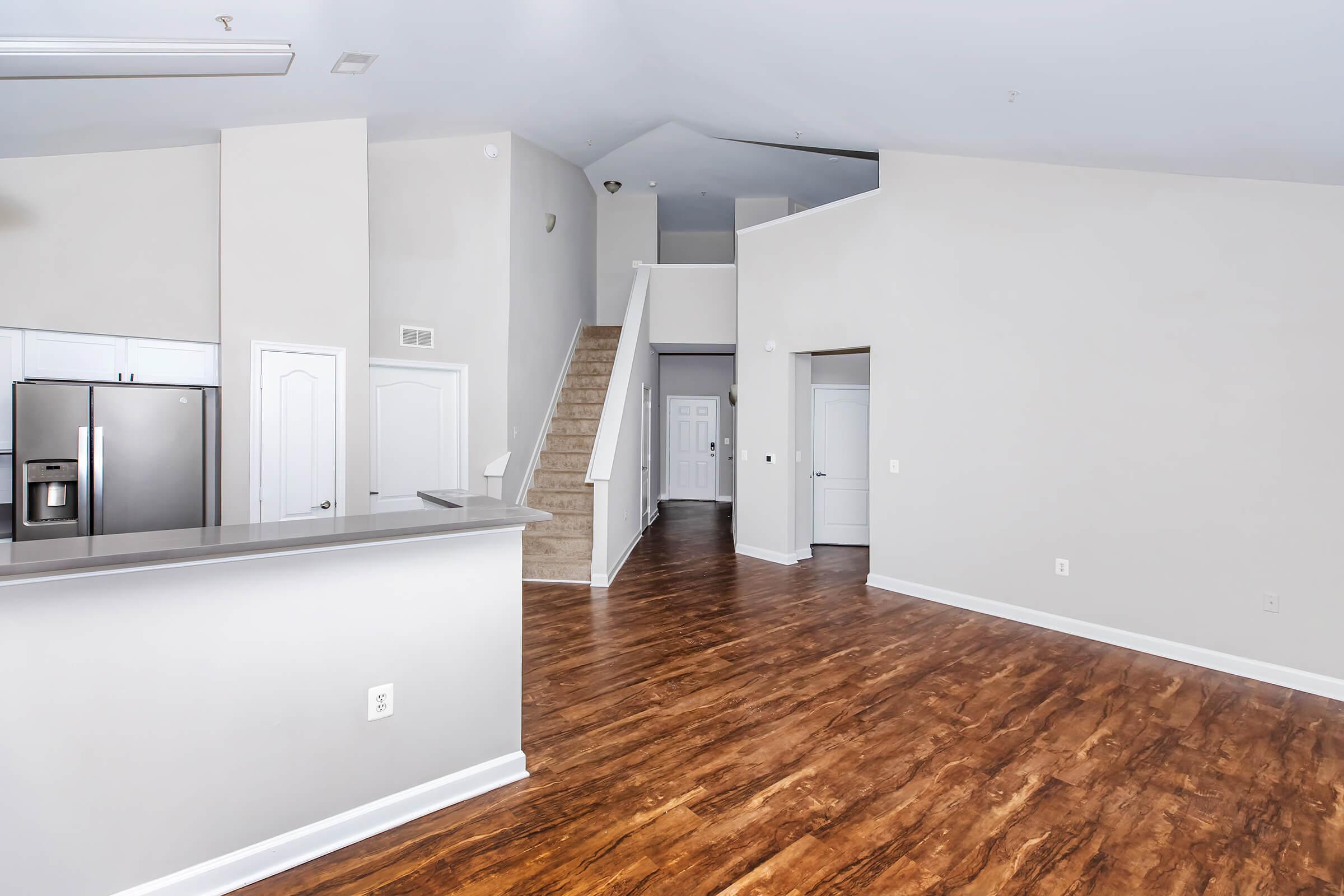 Spacious and modern interior of a residential living area featuring hardwood floors, a staircase leading to an upper level, and an open kitchen with stainless steel appliances. The walls are painted in neutral tones, creating a bright and inviting atmosphere.