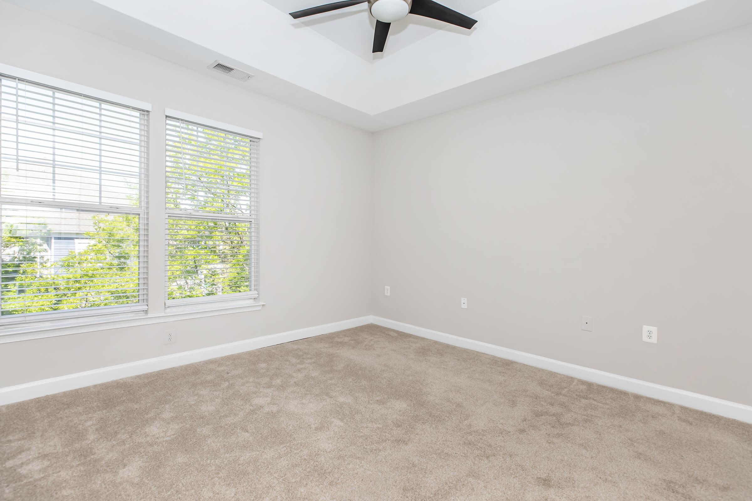 A spacious, empty room with light-colored walls and soft beige carpet. It features a ceiling fan and large windows with white blinds, allowing natural light to fill the space. The room is well-lit and appears ready for decoration or furnishing.