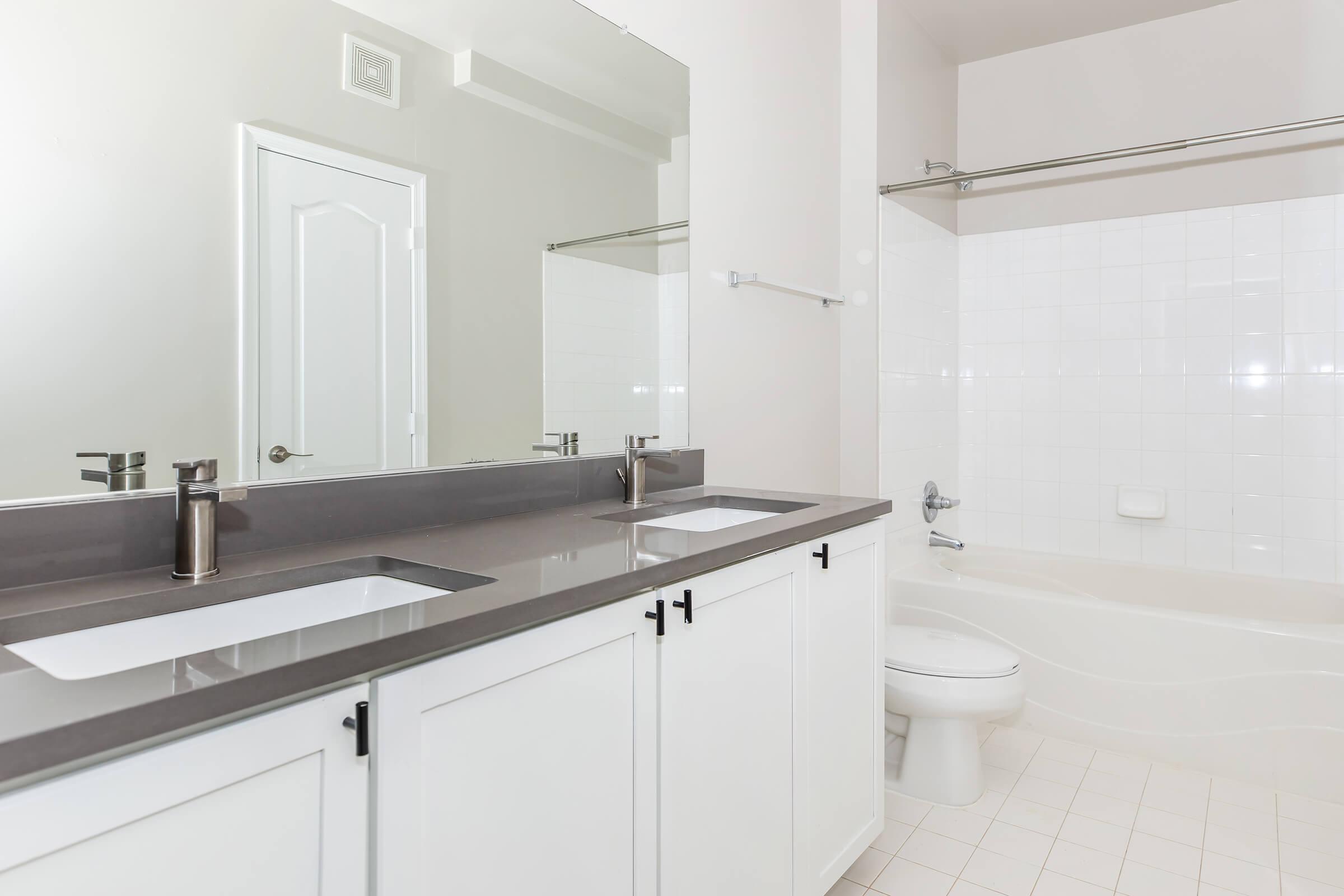 Modern bathroom featuring a double vanity with two sinks and sleek silver faucets, a large mirror above, and a light-colored tiled floor. The adjacent area includes a bathtub and a toilet. The walls are painted in a soft neutral color, creating a clean and spacious aesthetic.