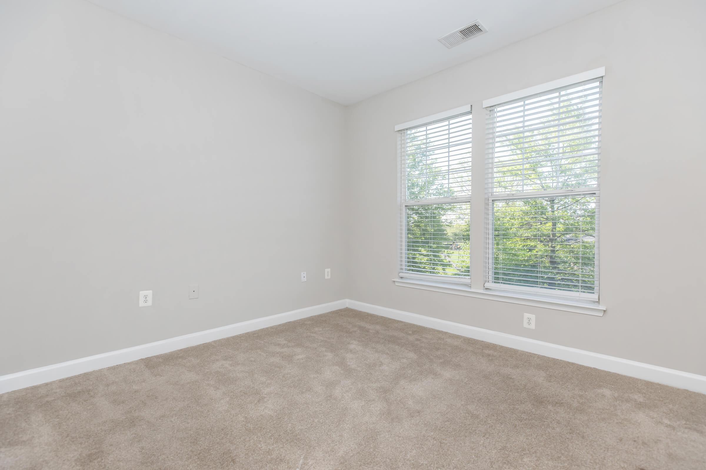 Empty room with light gray walls and beige carpet. Two windows with white blinds allow natural light to enter, revealing a view of greenery outside. The room is spacious with no furniture, providing a clean and minimalist aesthetic.
