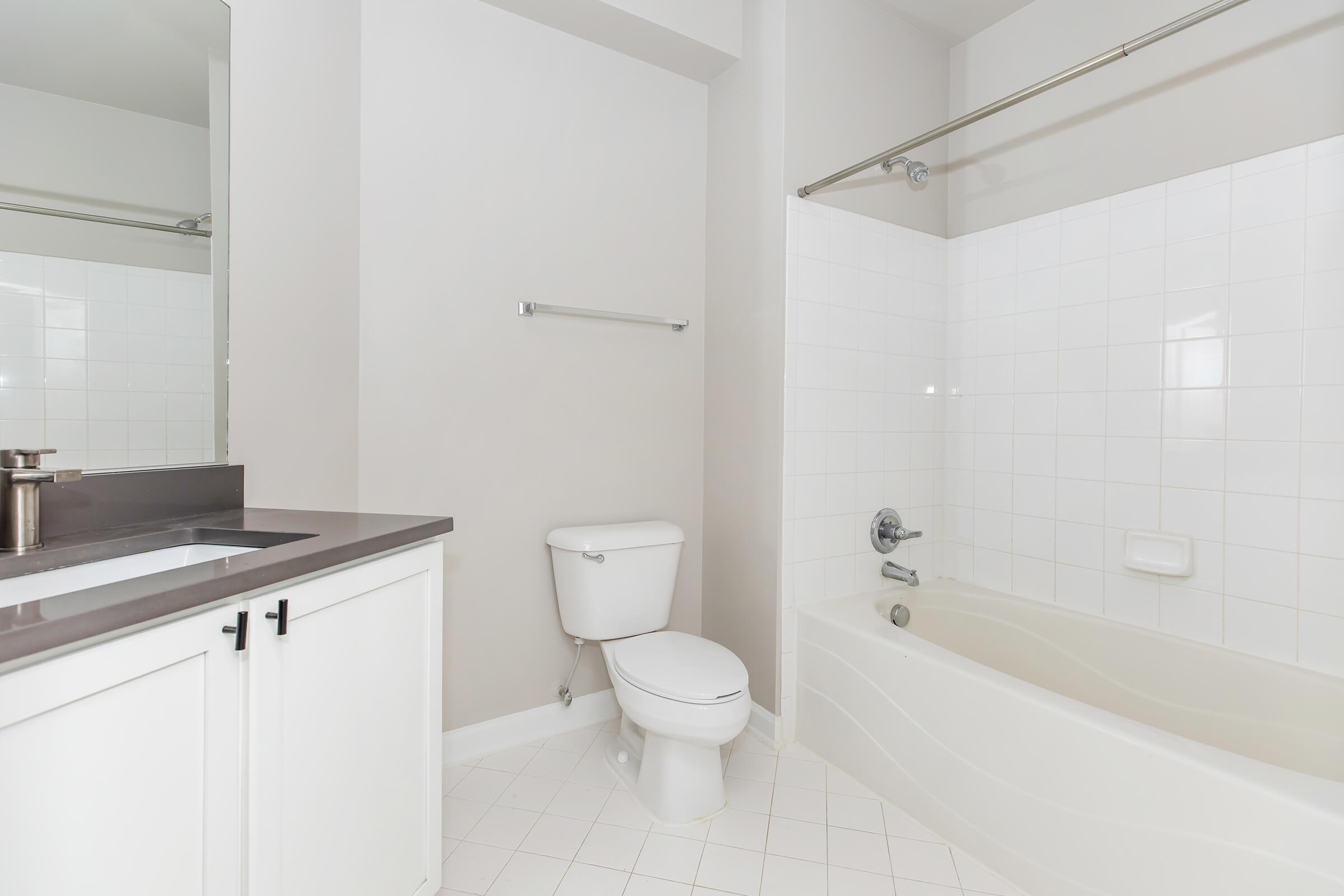 A clean and modern bathroom featuring a white bathtub, a shower curtain rod, a toilet, a sink with a dark countertop, and a large wall mirror. The walls are a light neutral color, and the flooring is tiled in a white pattern. The space is bright and uncluttered.