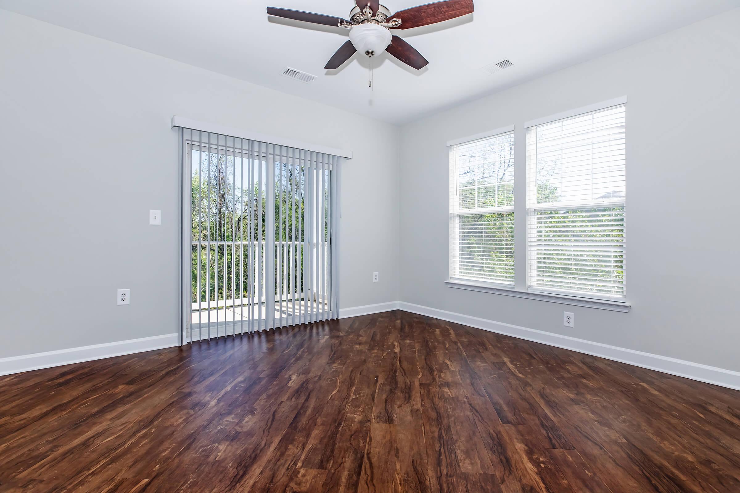 A spacious room with brown hardwood flooring, light gray walls, and a ceiling fan. Large windows provide natural light, and there is a sliding glass door leading to a balcony or patio area. Vertical blinds are partially drawn on the door, creating an open and airy atmosphere.