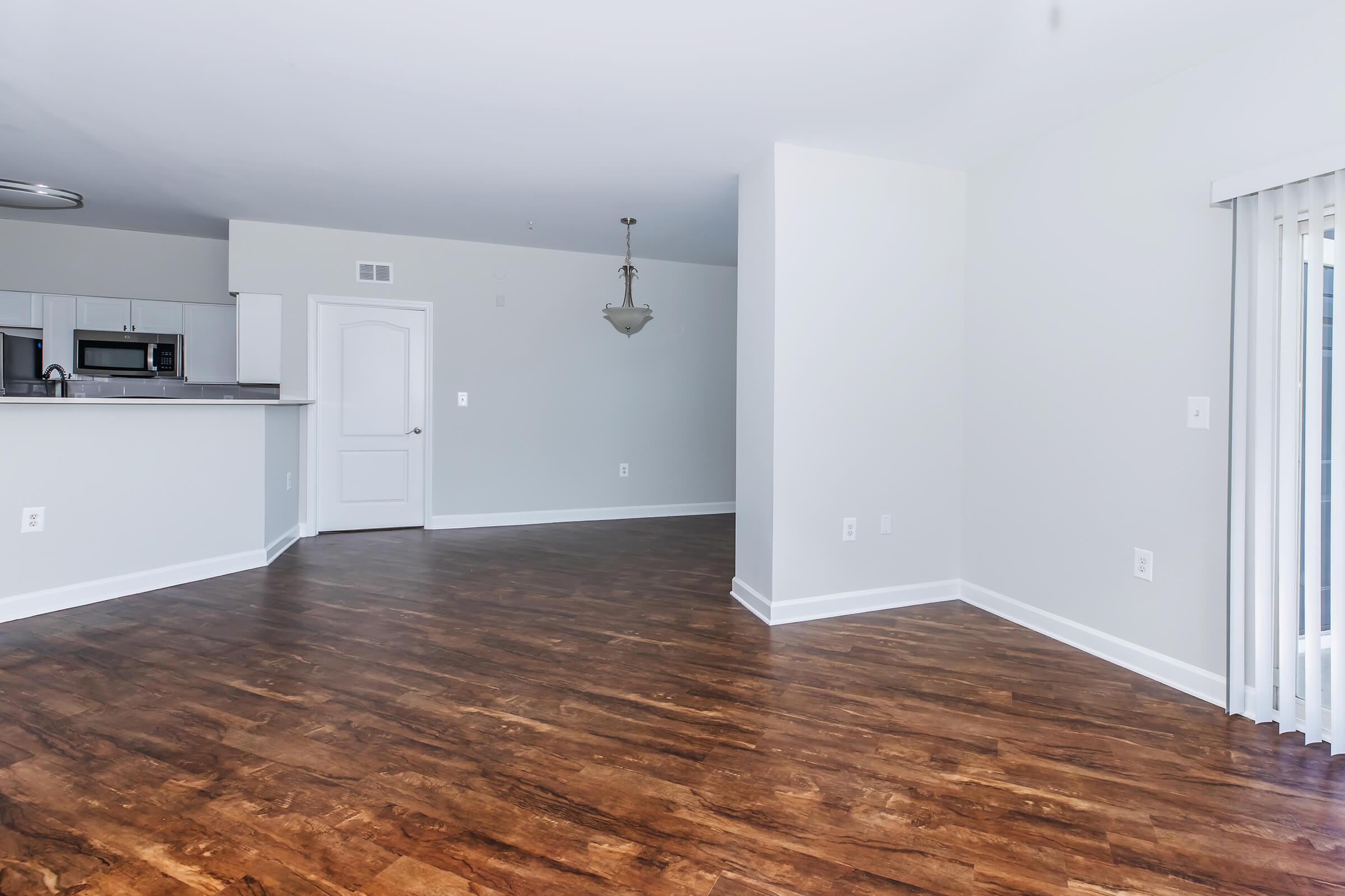 A spacious, empty living area featuring hardwood flooring, light-colored walls, and a single light fixture hanging from the ceiling. To the left, there's a view of a kitchen area with modern appliances visible. Large sliding glass doors open to a balcony, allowing natural light to fill the room.