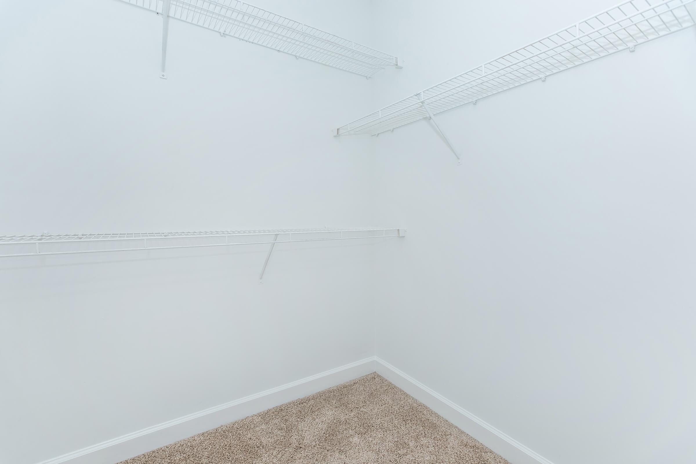 An empty closet with white walls and carpeted floor, featuring three wire shelving units mounted on the walls, offering storage space. The overall look is clean and minimalist, with no items present.